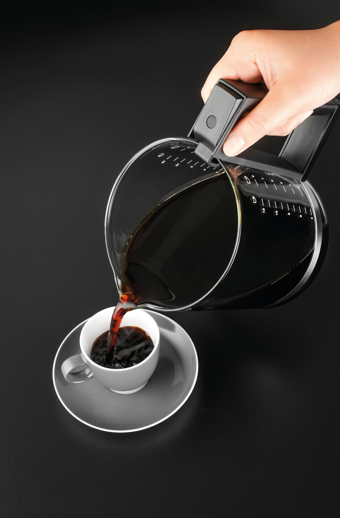 Osoba nalewa kawę z karafki do białego kubka na szarej spodku. Czarne tło.