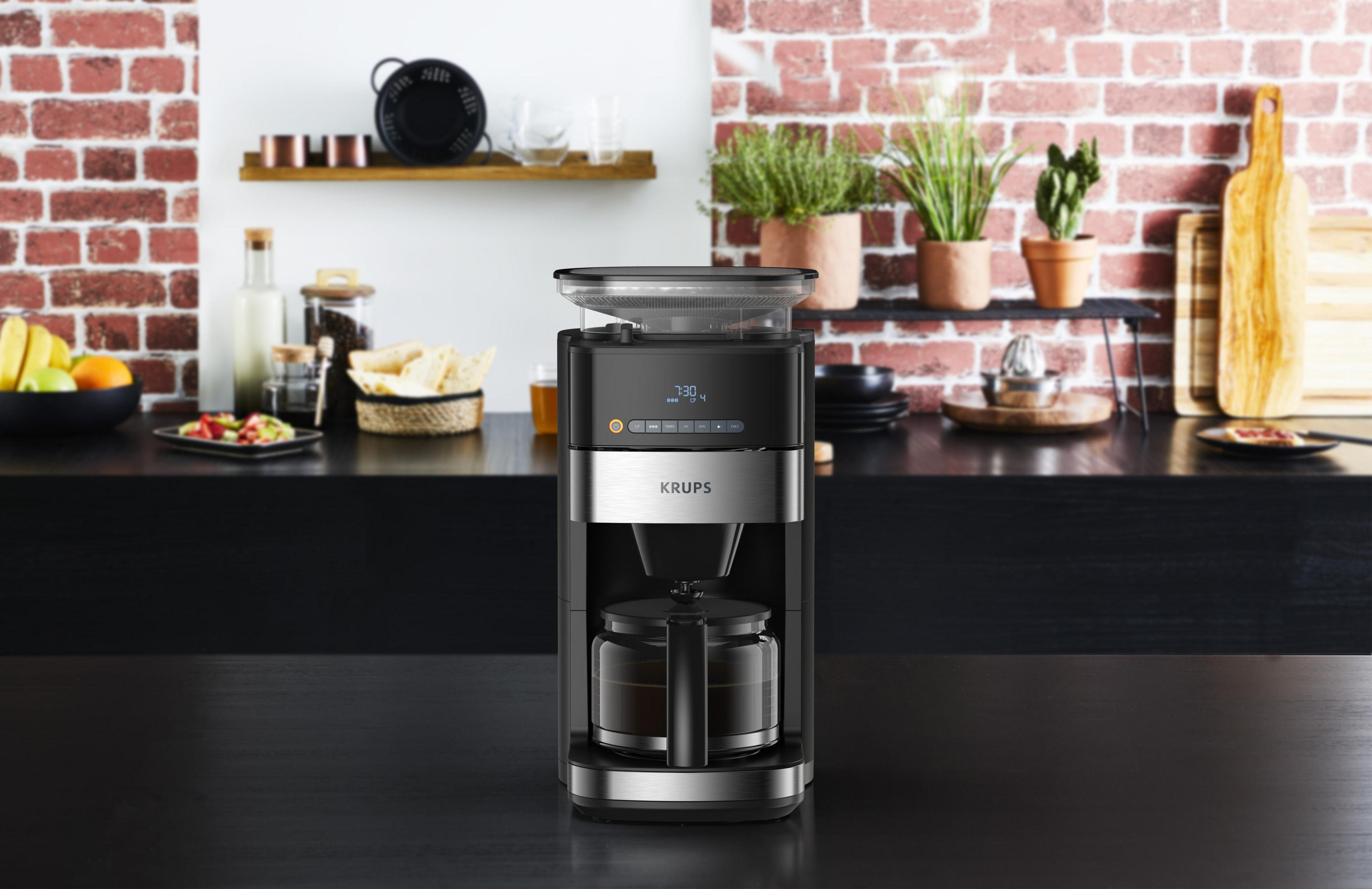 Czarny i srebrny ekspres do kawy KRUPS na blacie kuchennym. Tło ma ceglaną ścianę i półki z przedmiotami.