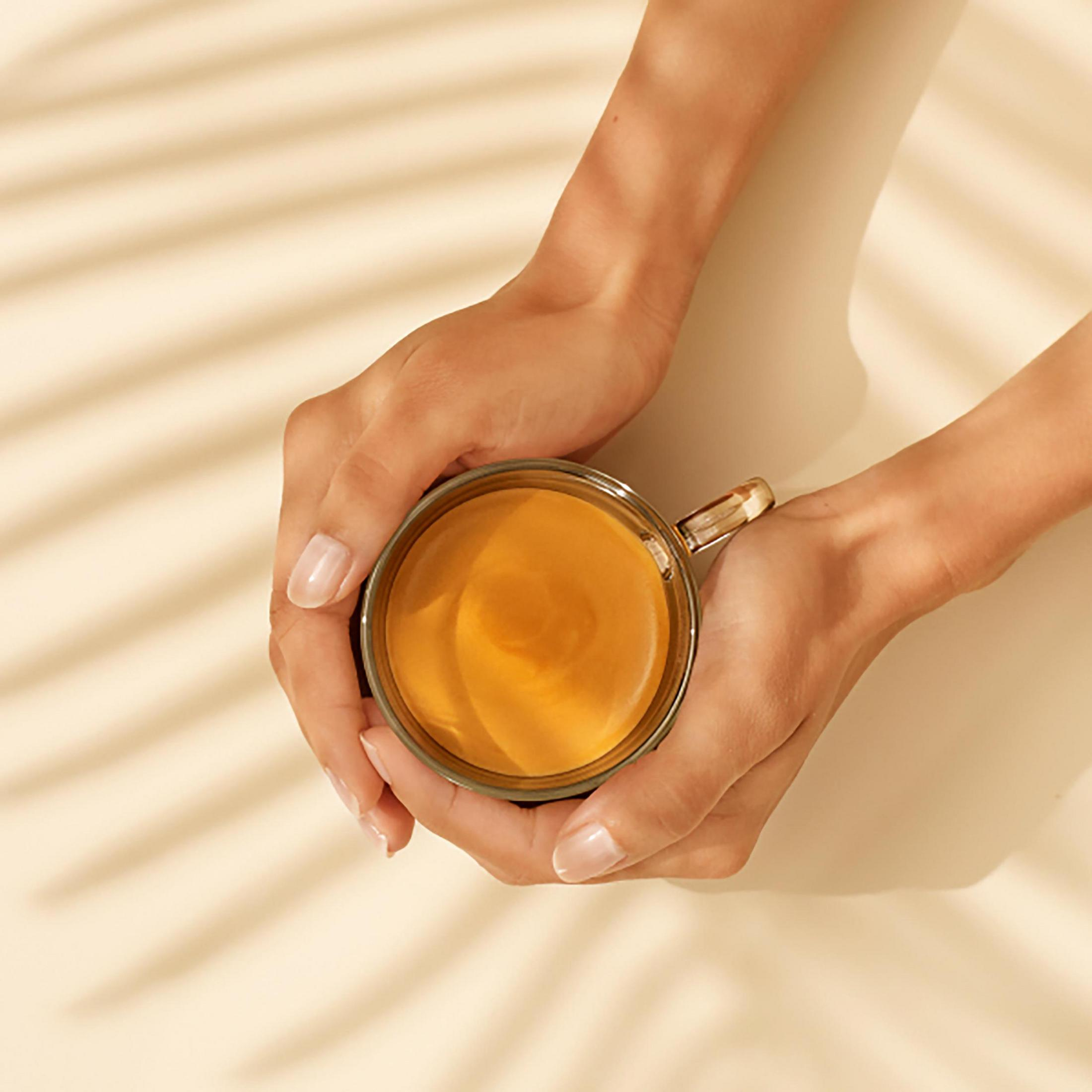 Dłonie trzymają szklany kubek kawy. Dłonie mają jasną karnację. Cienie palm są widoczne w tle.