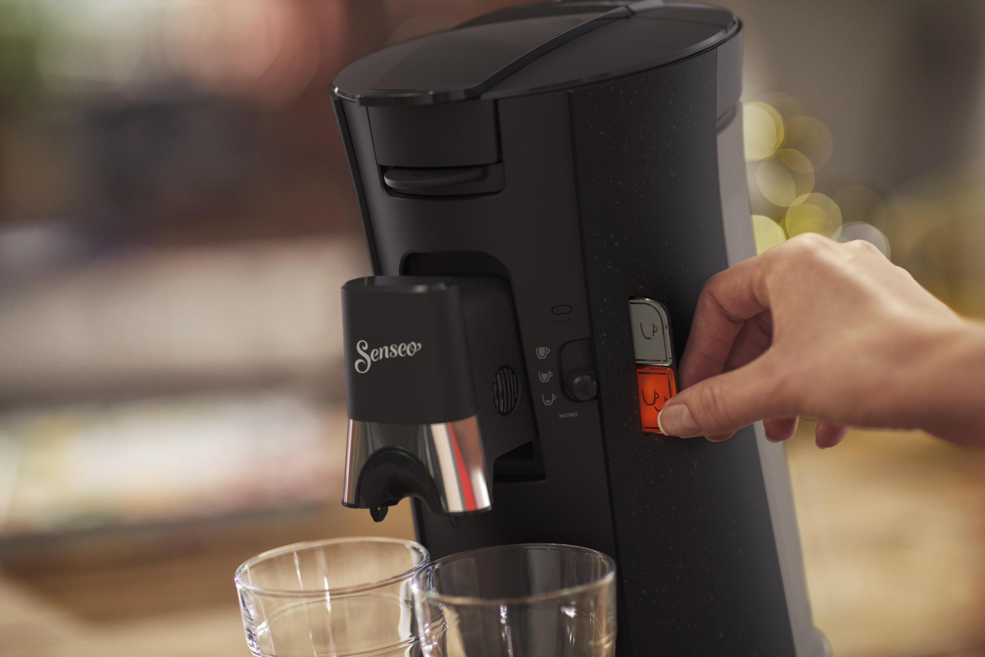 Schwarze Senseo-Kaffeemaschine, mit einer Hand, die einen orangenen Knopf drückt. Zwei Gläser stehen davor.