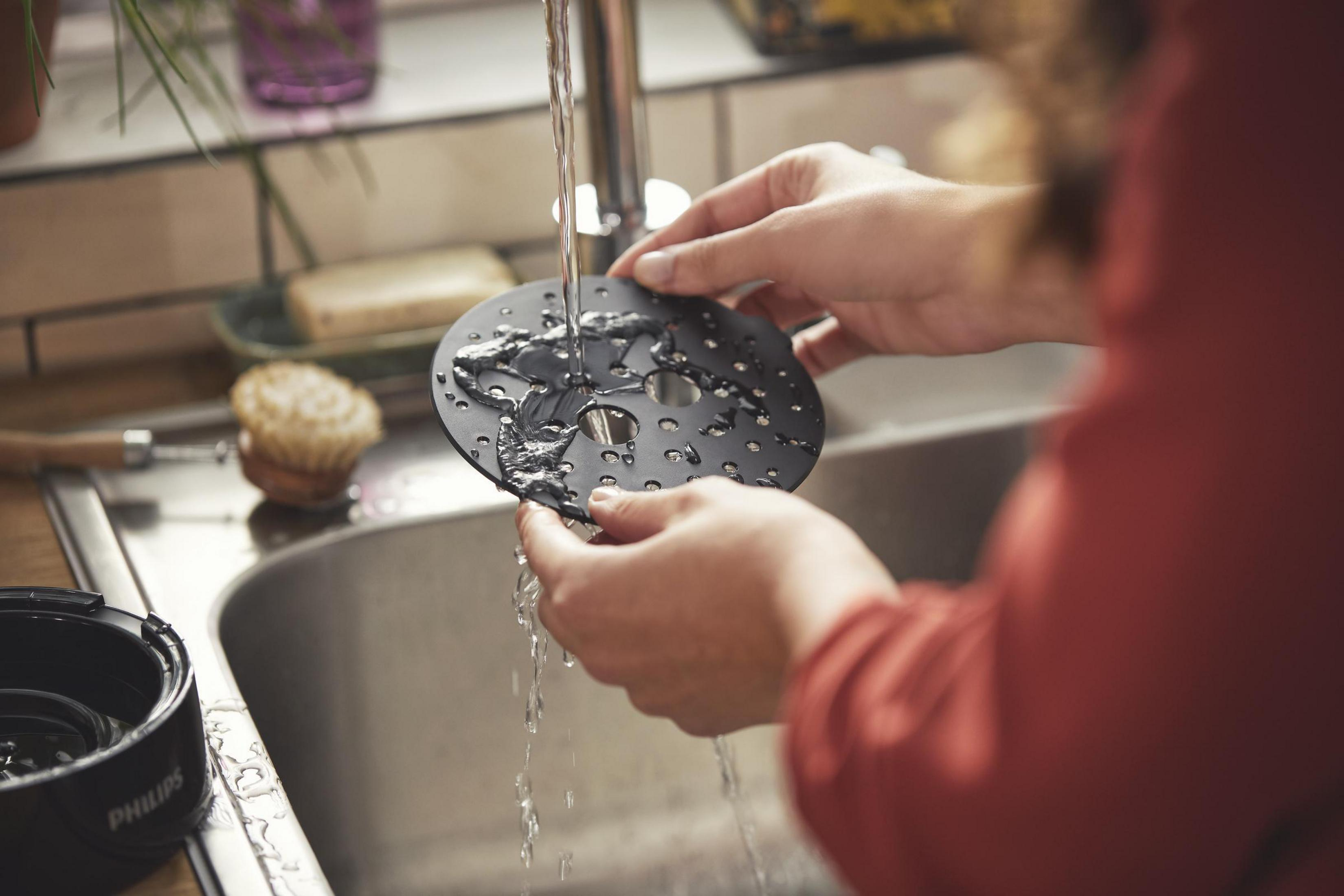 Eine Person wäscht ein schwarzes Küchengerät unter fließendem Wasser in einem Edelstahlbecken. Eine Bürste und Seife liegen daneben.
