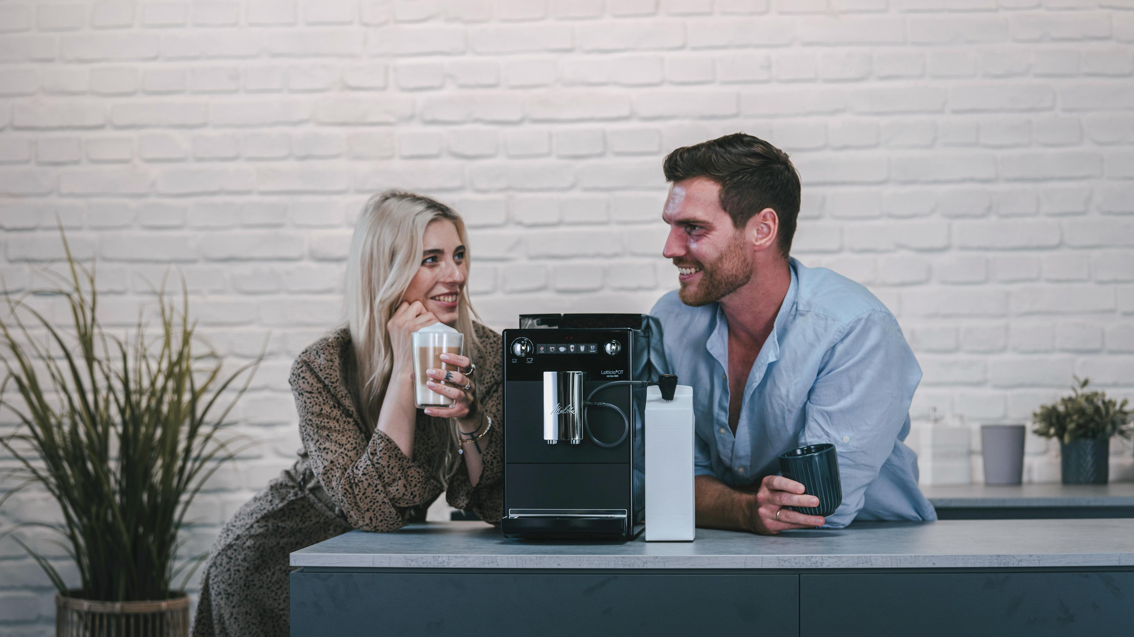 Ein Paar an einer Kaffeemaschine. Die Frau hält einen Latte, der Mann einen Becher, sie lächeln.