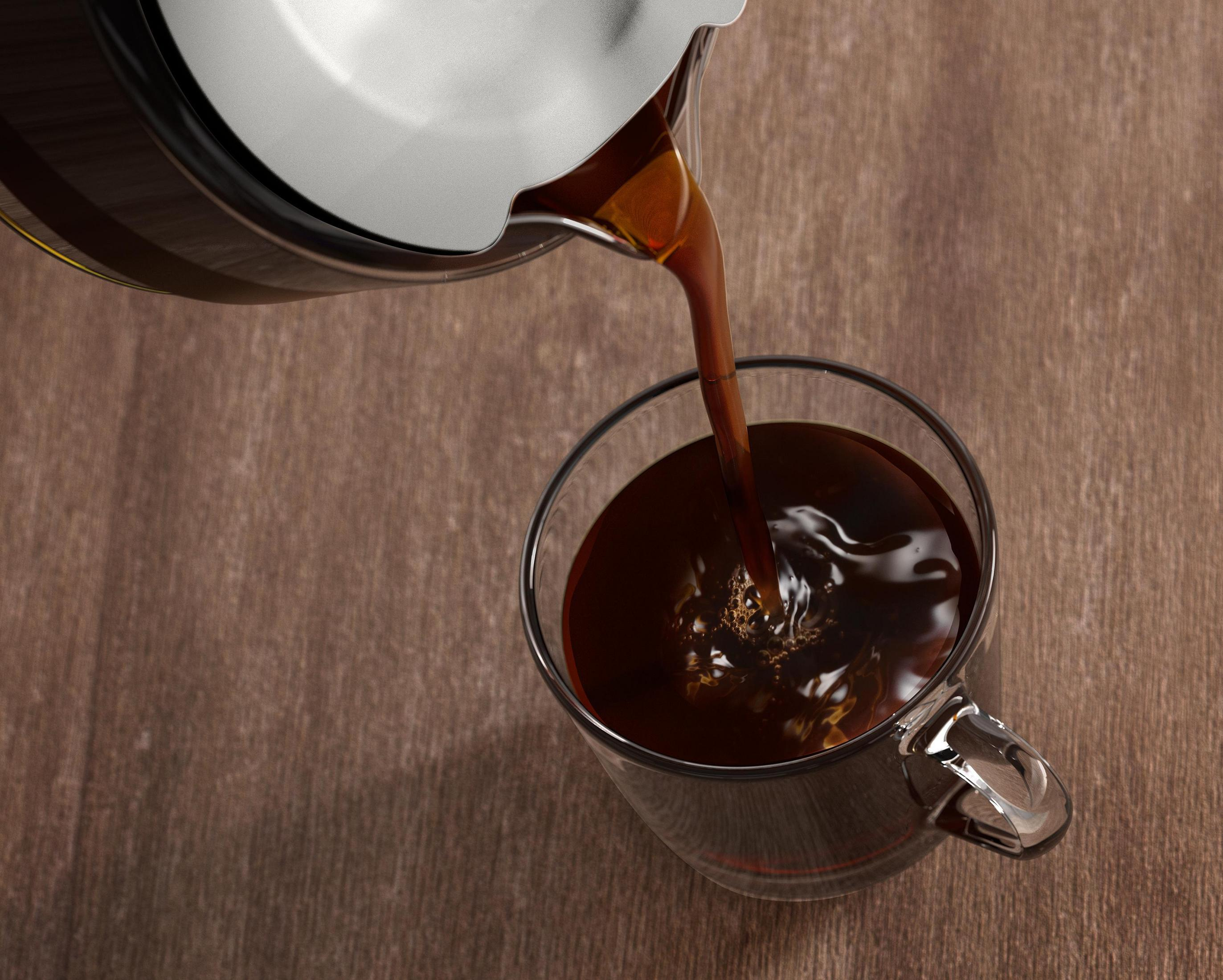 Kawa jest wlewana ze srebrnego dzbanka do szklanego kubka na drewnianym stole. Kawa jest ciemna i wypełnia szklankę.