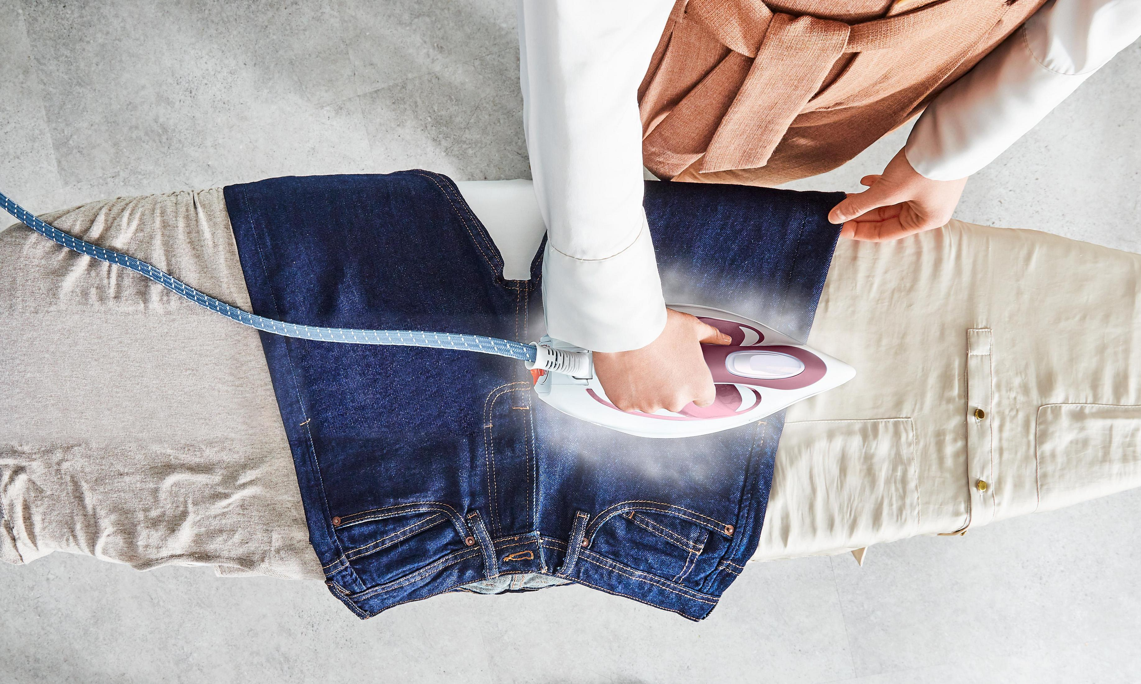 Osoba prasuje niebieskie dżinsy na desce do prasowania, z szarą i beżową tkaniną.