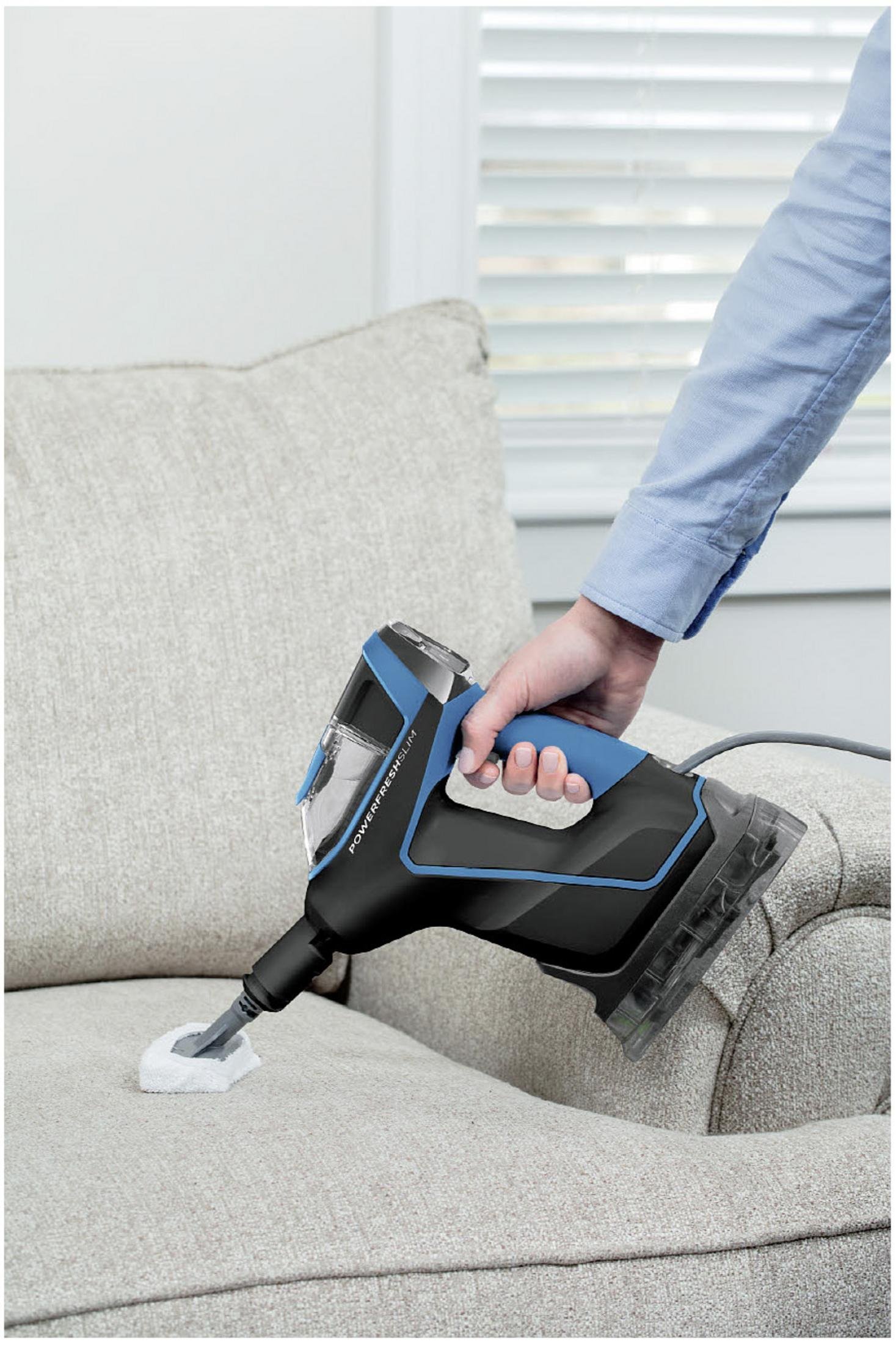 Osoba czyszcząca beżową sofę niebiesko-czarnym odkurzaczem parowym ręcznym.