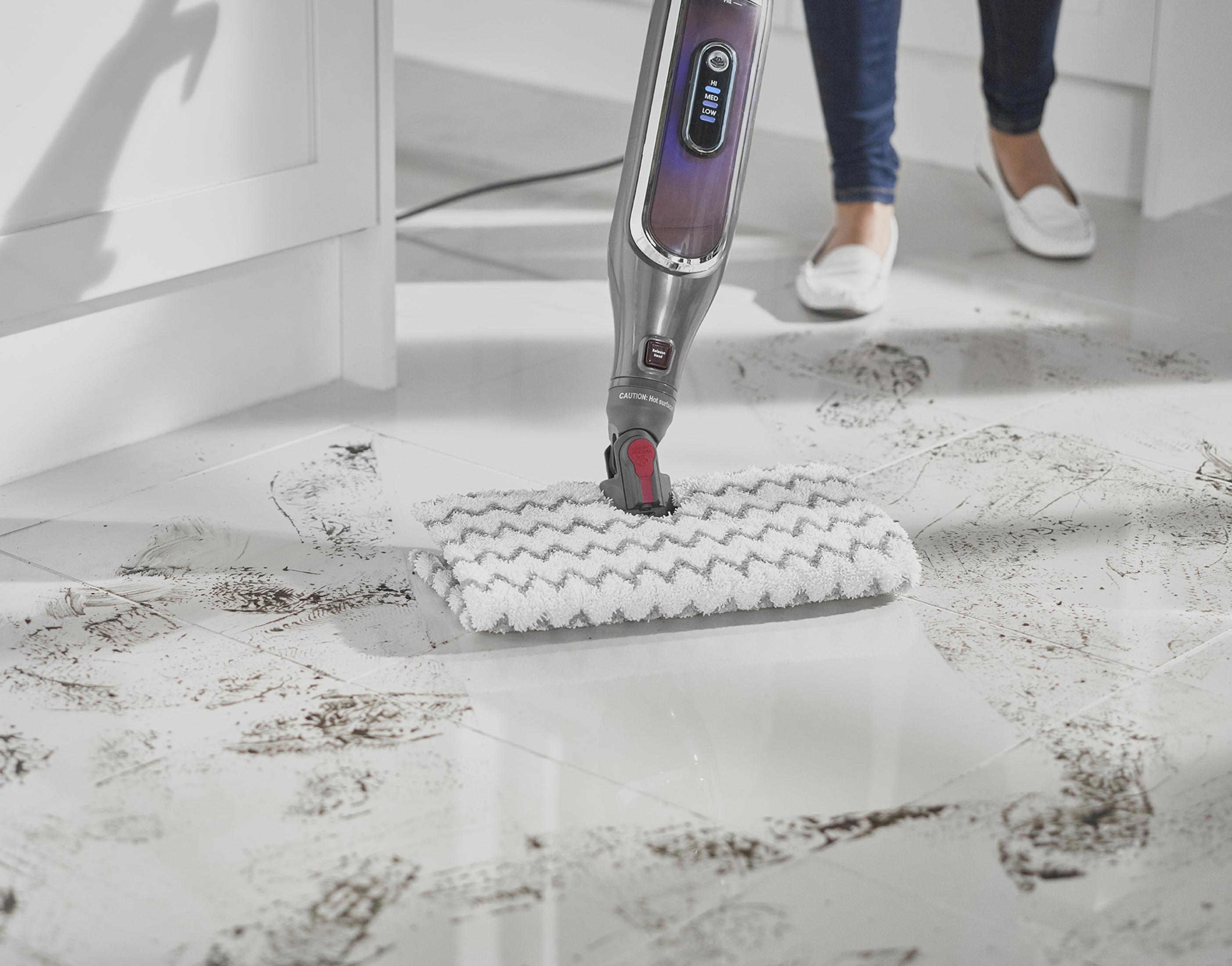 Kobieta czyści białą podłogę z płytek mopem parowym. Podłoga jest brudna. Mop jest ciemnoszary i czerwony.