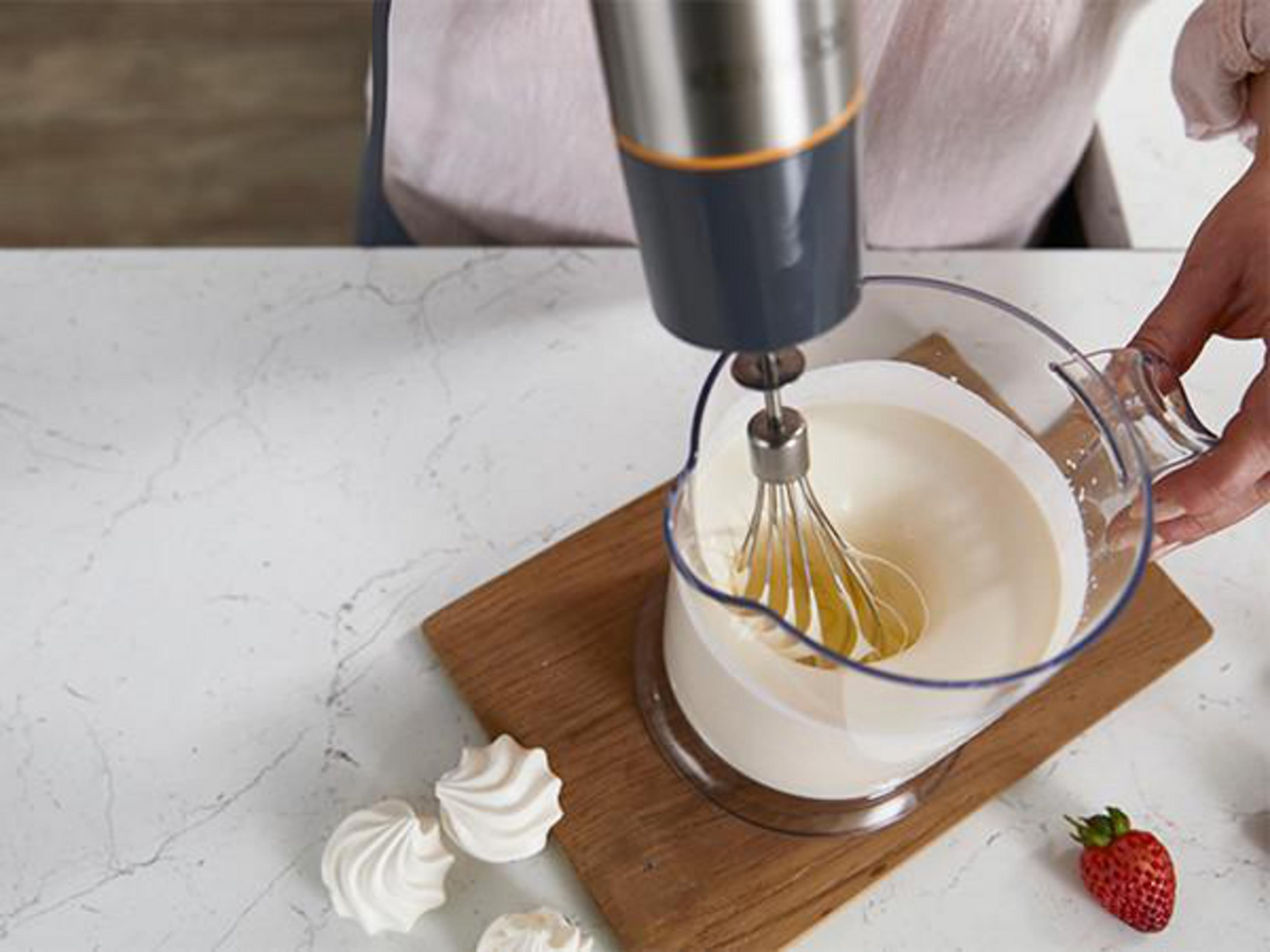 Eine Person, die einen Handmixer verwendet, um Sahne in einem durchsichtigen Krug aufzuschlagen. Erdbeeren und Baiser.