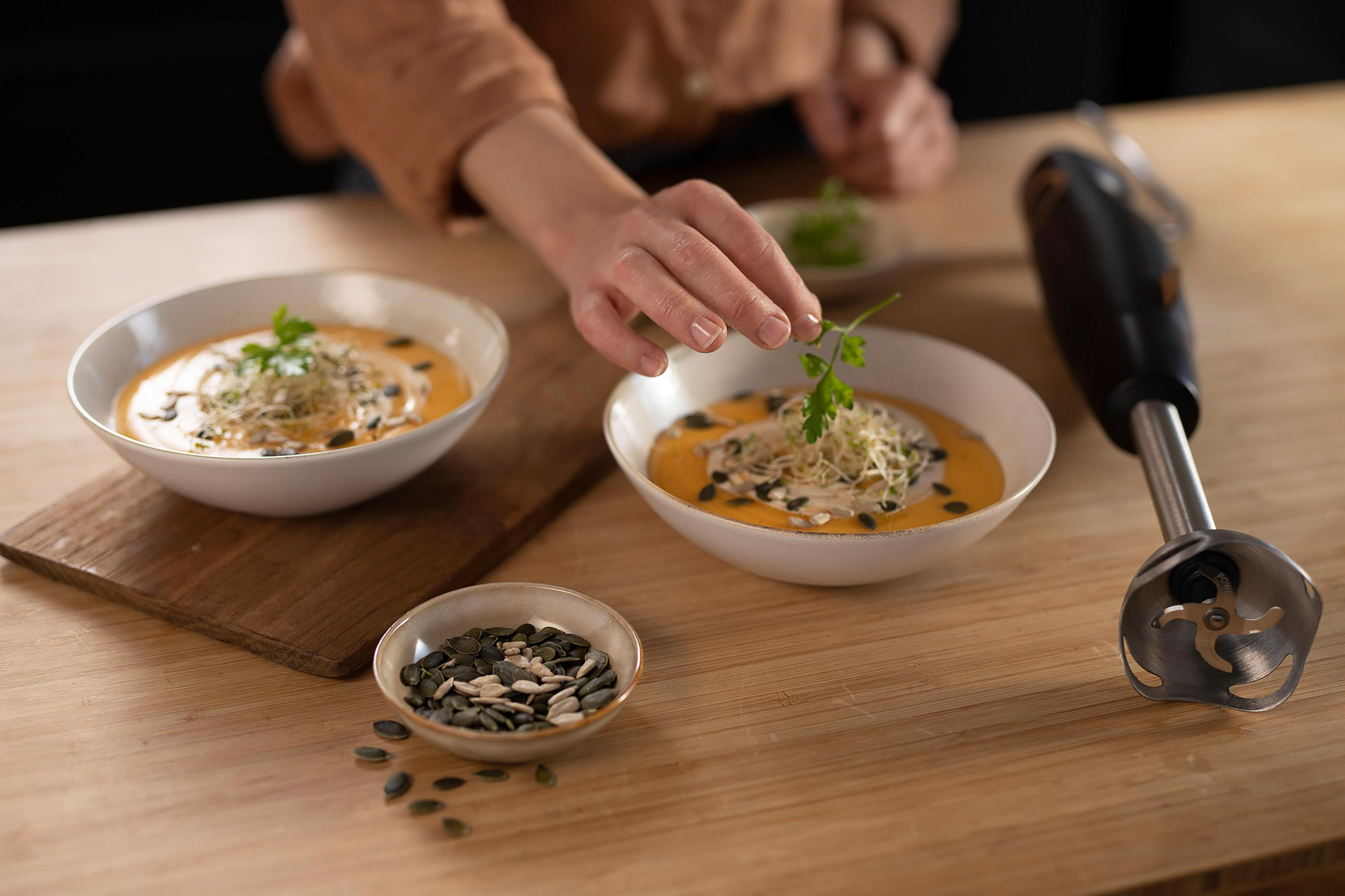 Due ciotole di zuppa con erbe aromatiche, semi di zucca e un frullatore a immersione su un tavolo di legno.