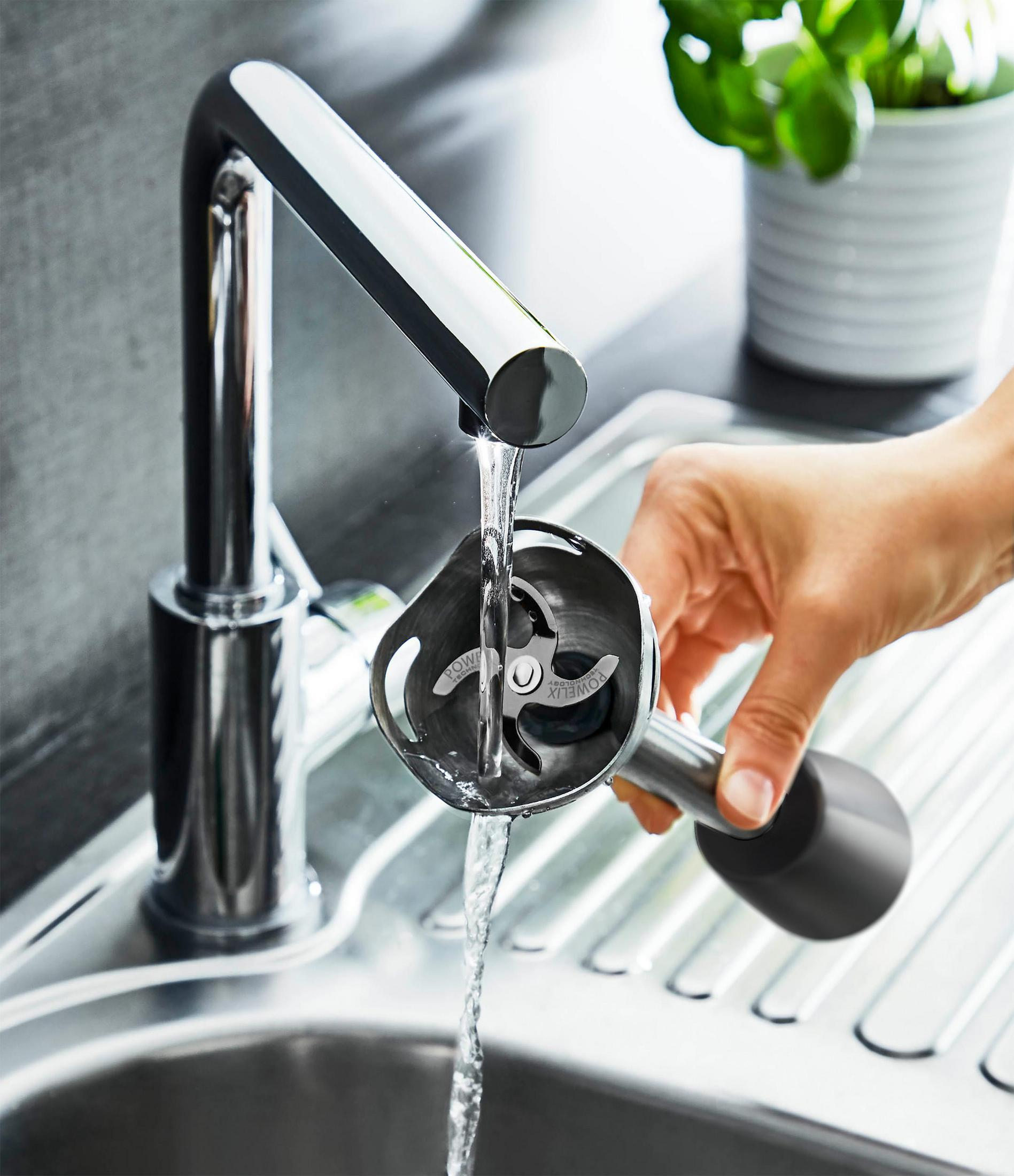 Una mano risciacqua un accessorio argentato per frullatore a immersione sotto un rubinetto cromato.