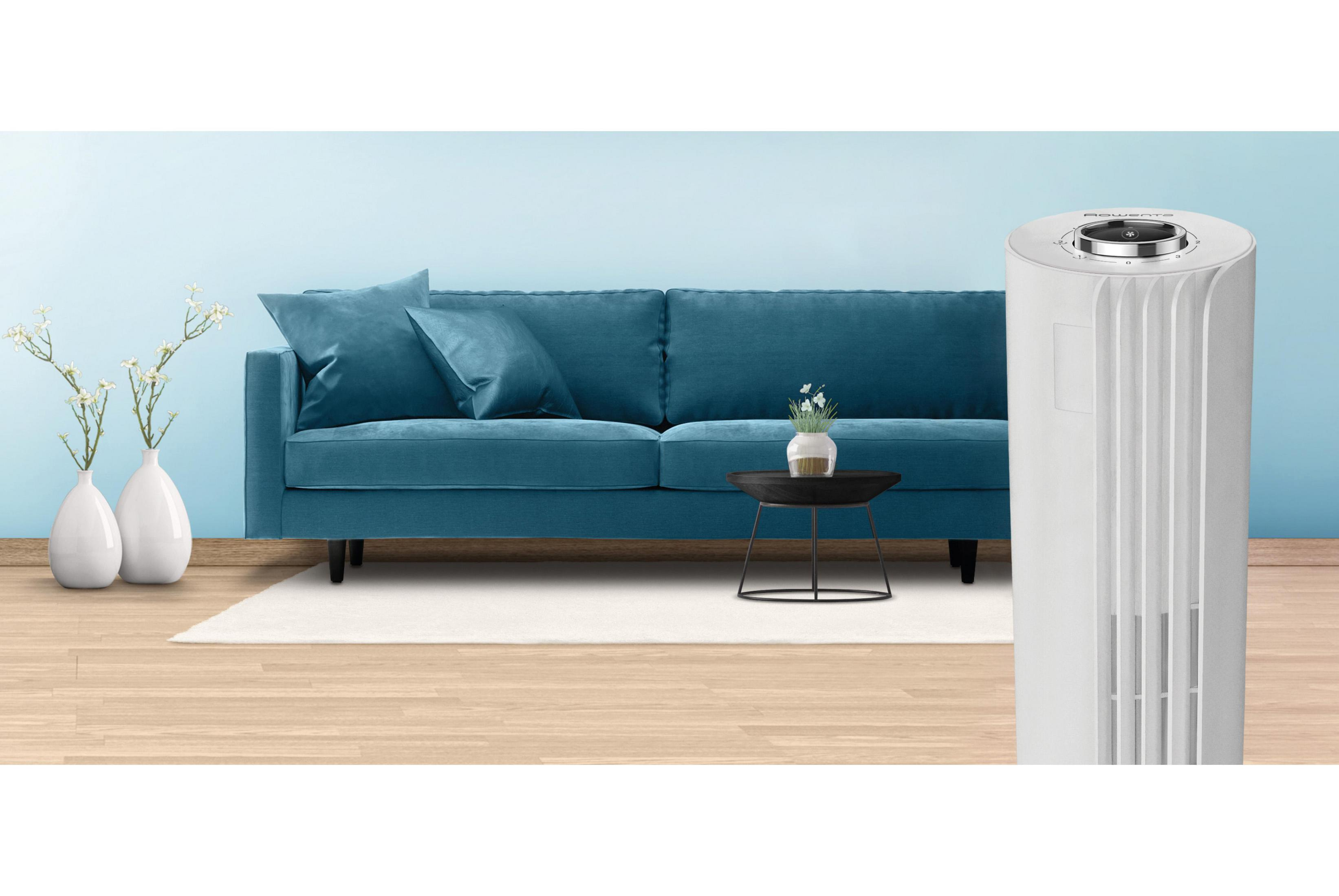 Un purificador de aire blanco junto a un sofá azul con almohadas. Una mesa tiene flores.