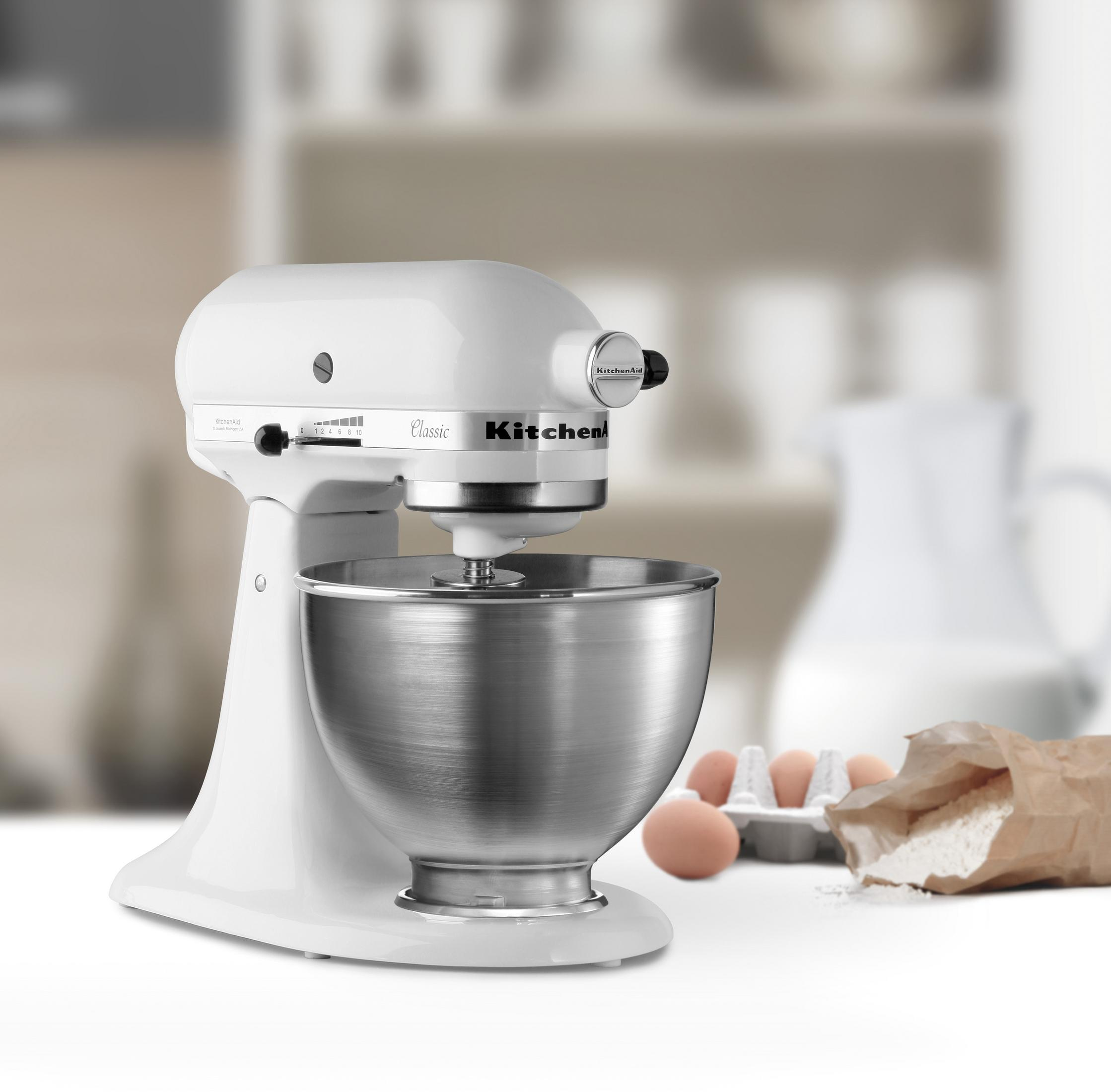Weißer KitchenAid-Mixer mit silberner Schüssel, Eiern, Mehlbeutel und Krug im Hintergrund.