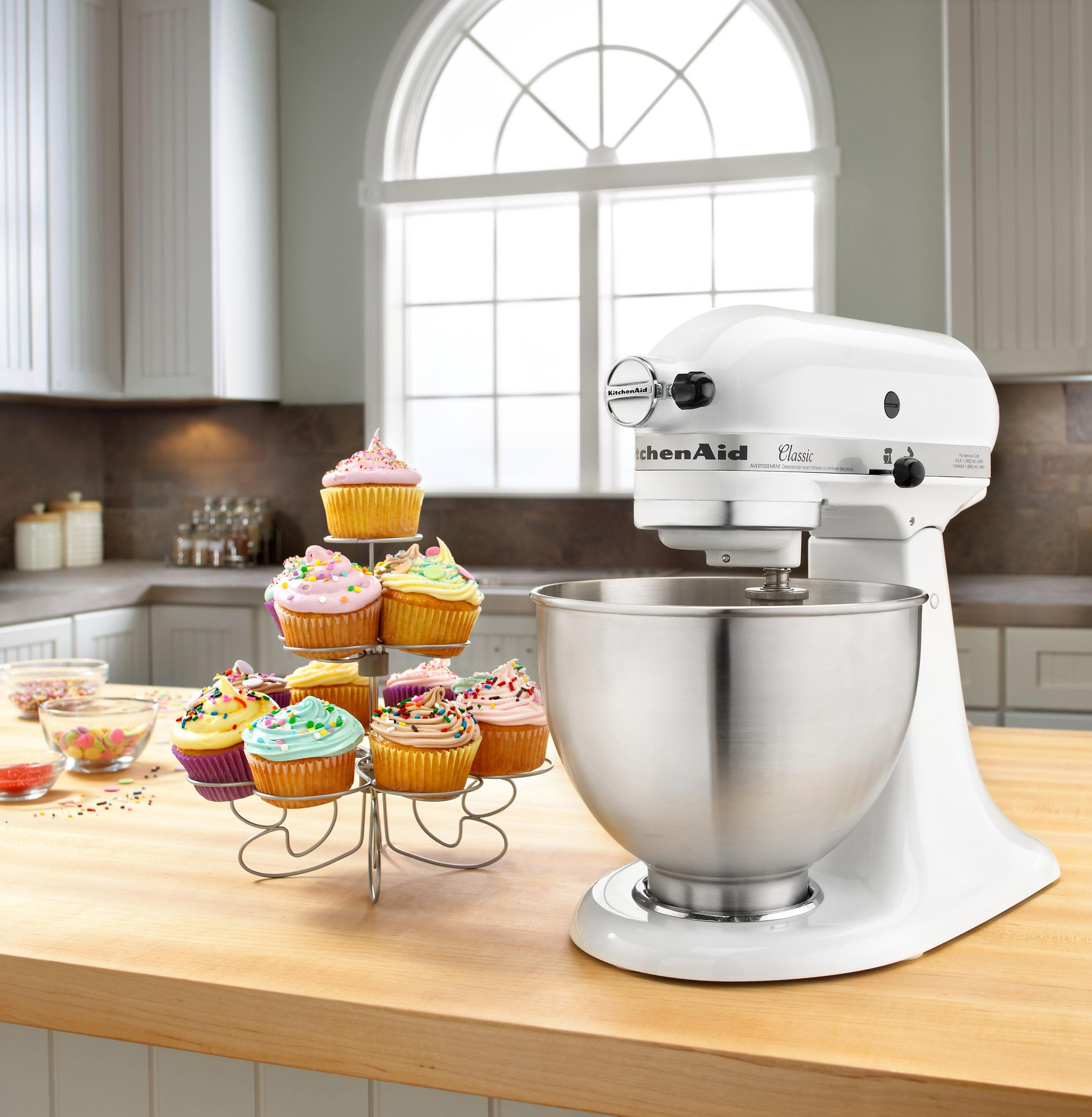 Weiße KitchenAid Küchenmaschine. Cupcakes auf einem Ständer. Holztheke und Küche im Hintergrund.