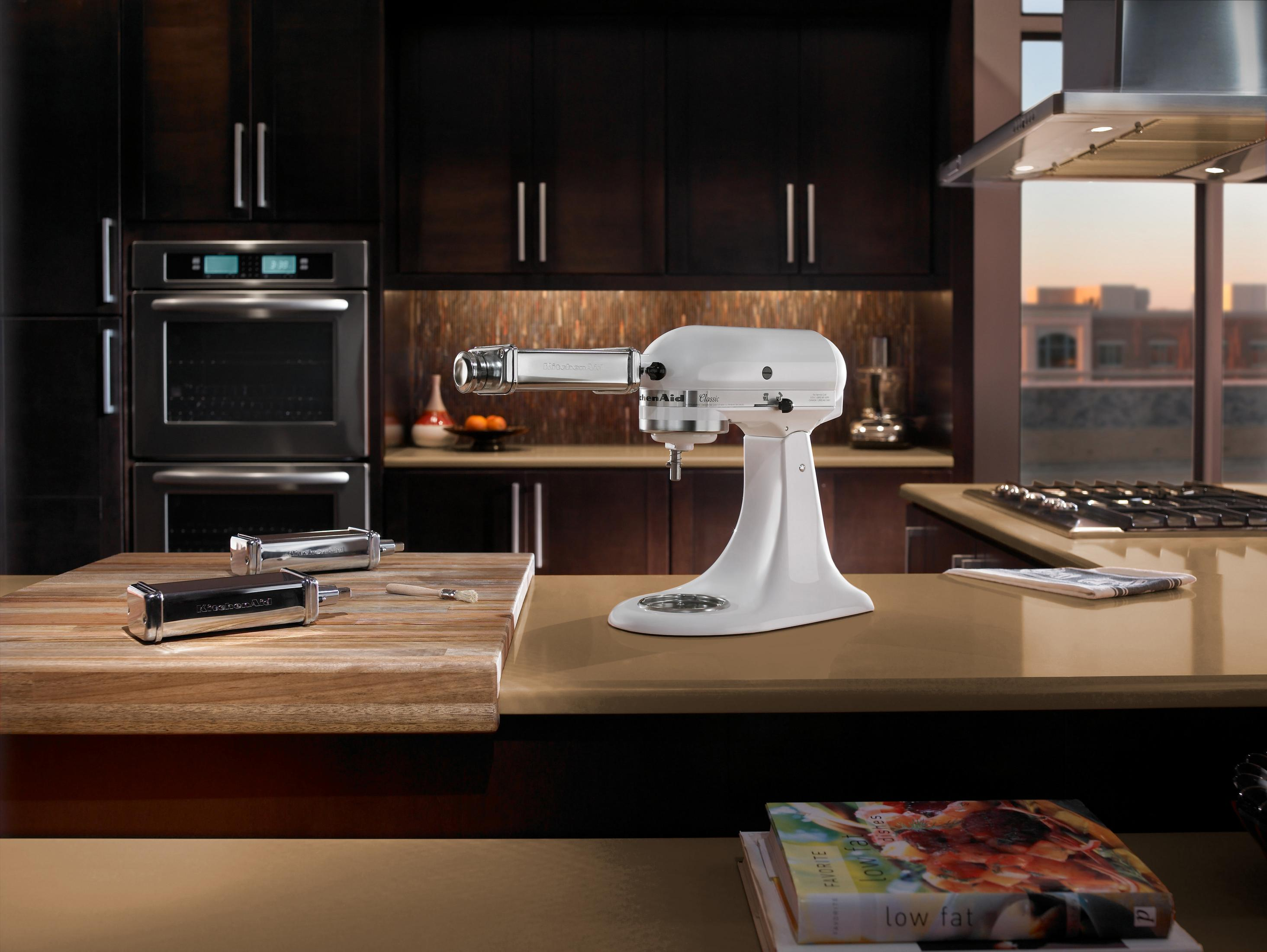 Ein weißer KitchenAid-Küchenmixer mit Aufsätzen auf einer Holztheke in einer Küche. Dunkle Schränke.