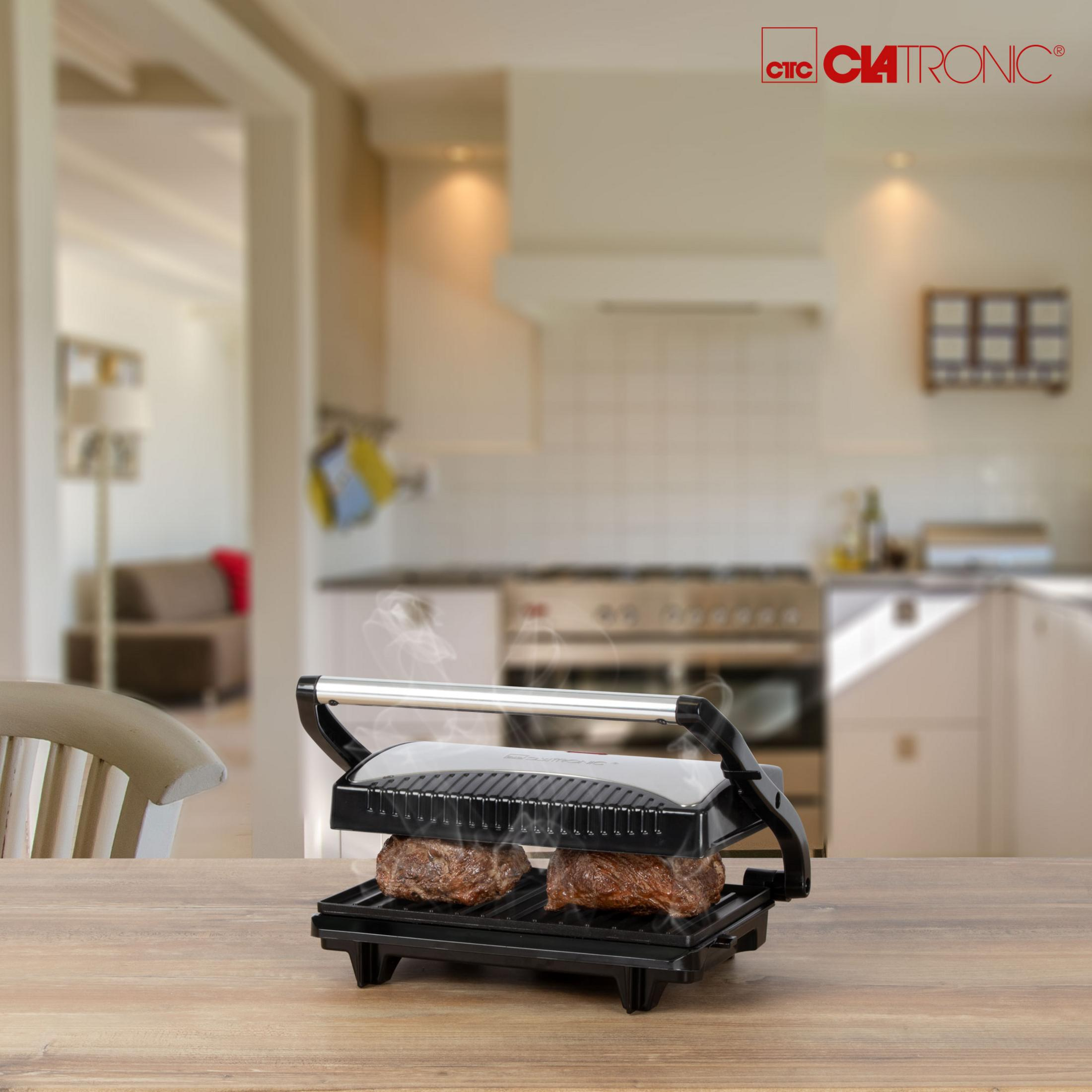 Essen wird in einem Sandwichmaker auf einem Holztisch gegrillt. Küche und Wohnzimmer im Hintergrund.