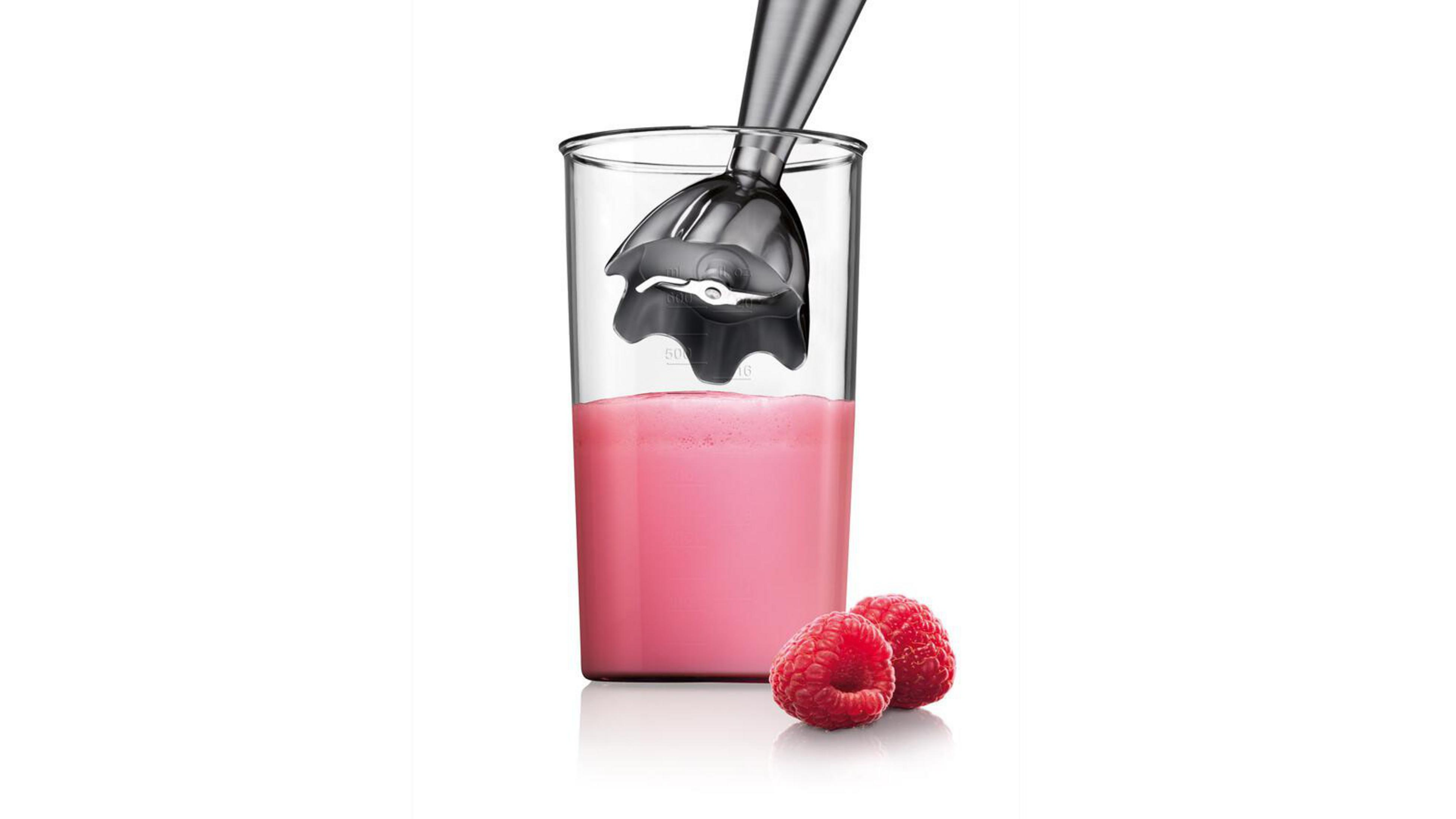 Blender ręczny w szklance z różowym płynem. Dwa maliny na białej powierzchni.