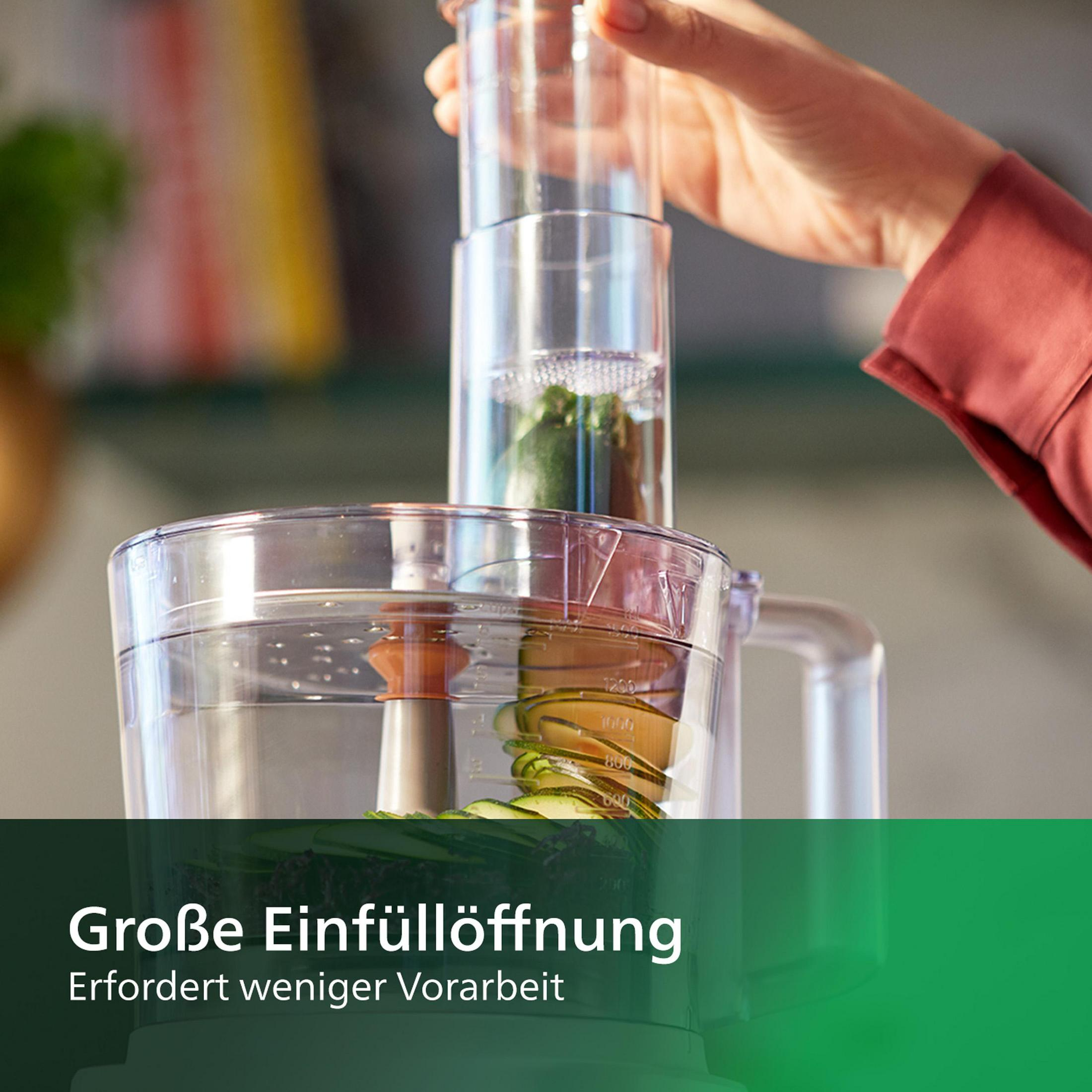 A food processor. Hand is placing zucchini into the top. Green background.