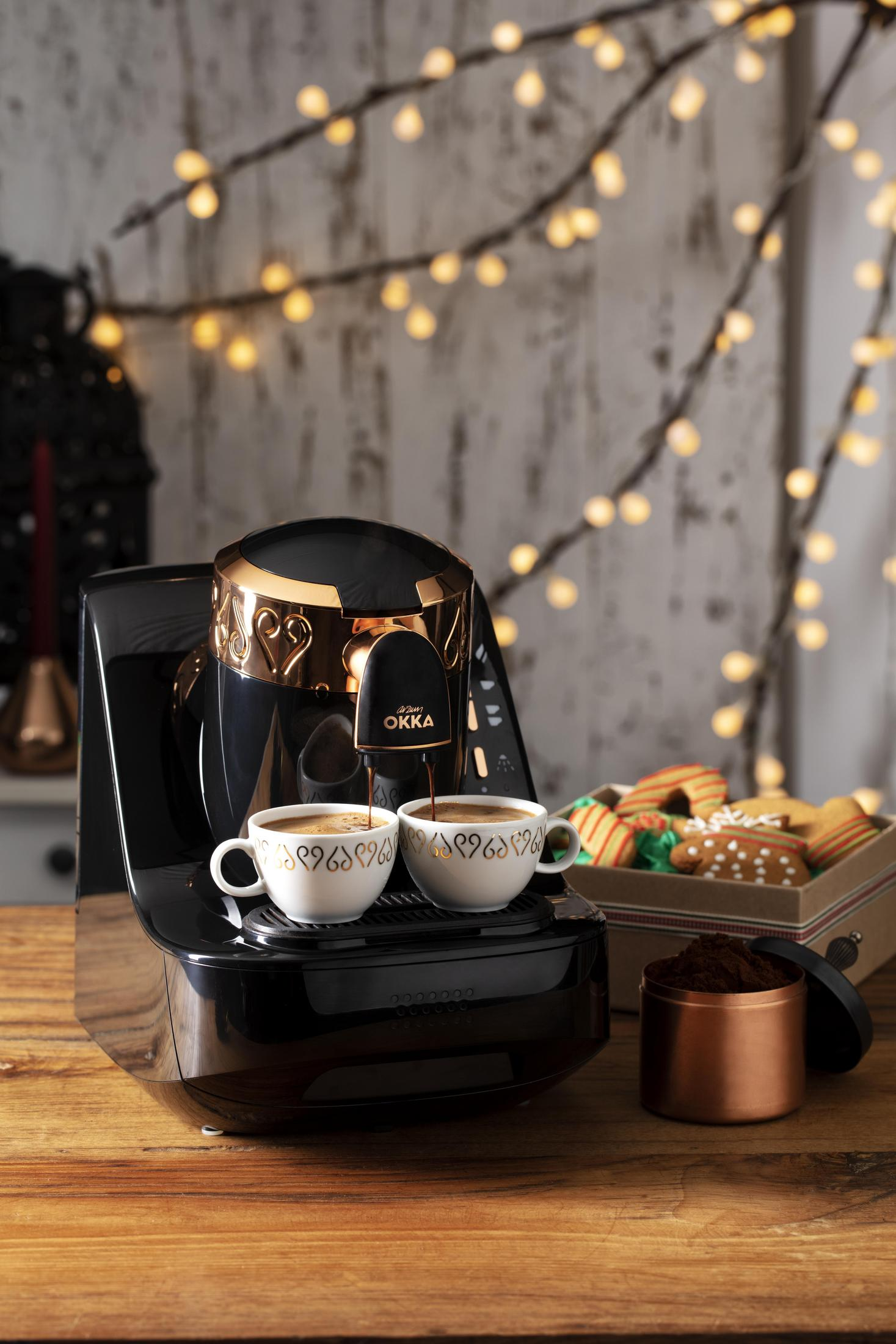 Kaffeemaschine gießt Kaffee in zwei Tassen. Kekse und Lichter im Hintergrund.