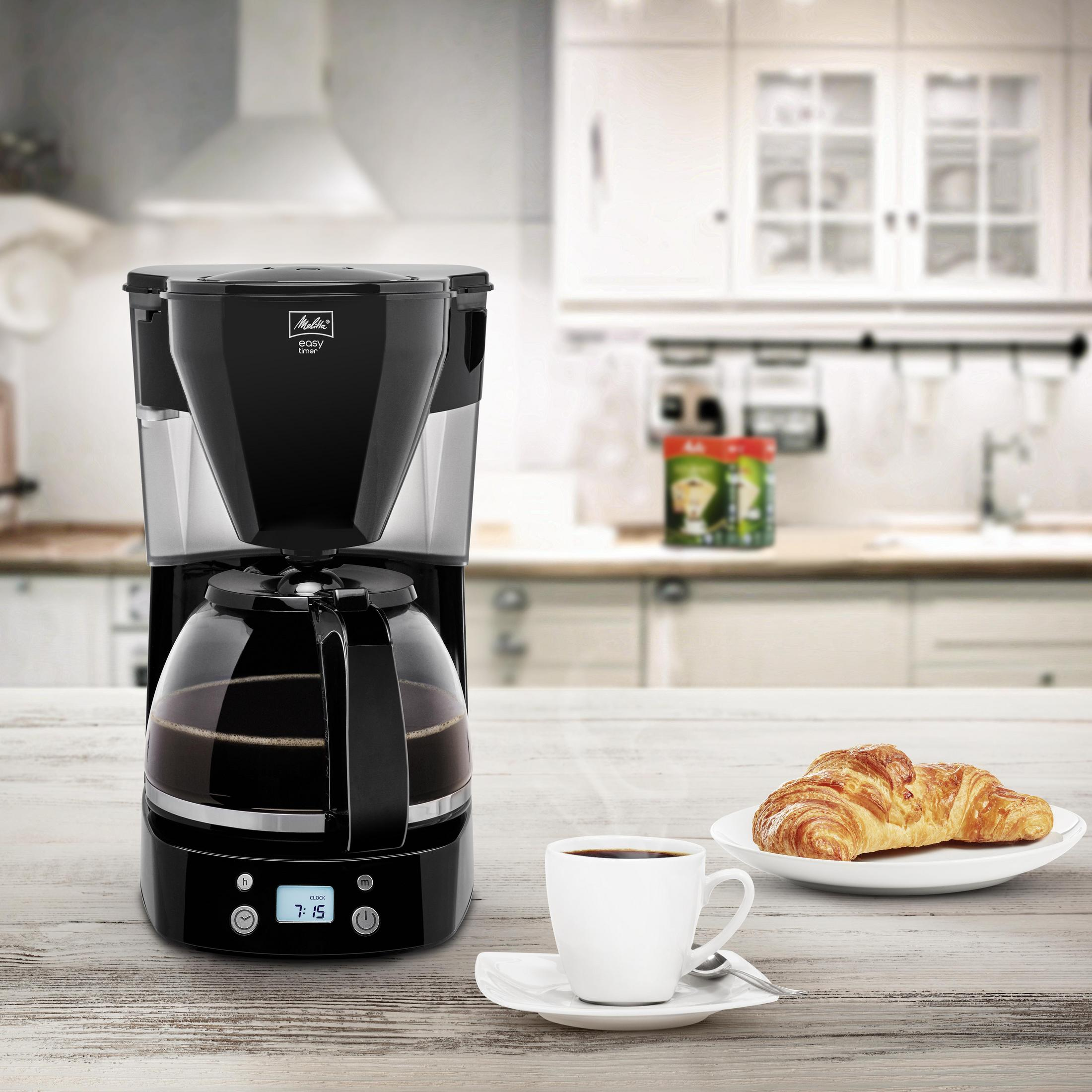 Czarny ekspres do kawy z pełną karafką, filiżanką kawy i rogalikiem.