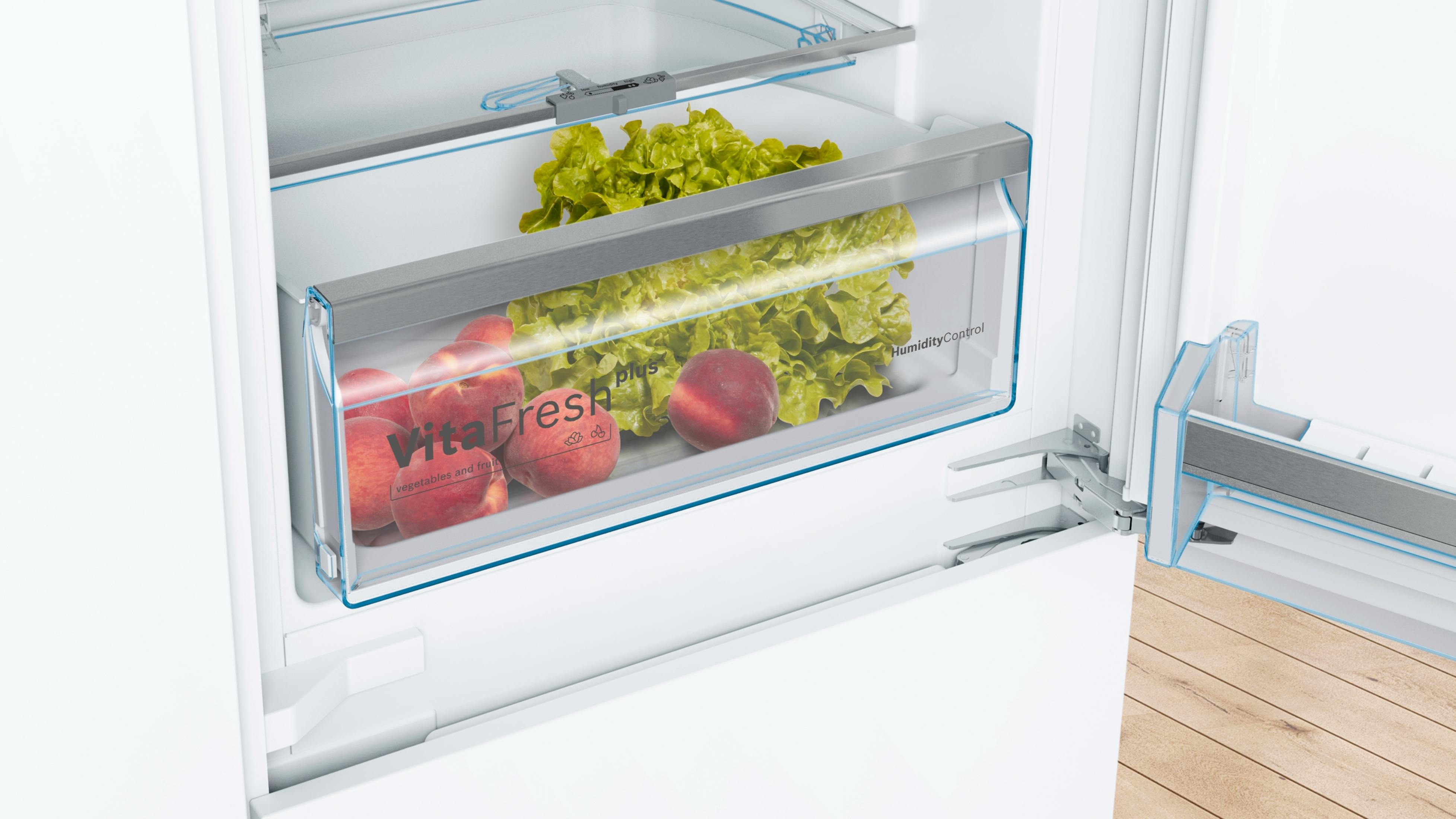 Interior de un refrigerador con frutas y verduras en un recipiente transparente. Puerta de nevera abierta visible.