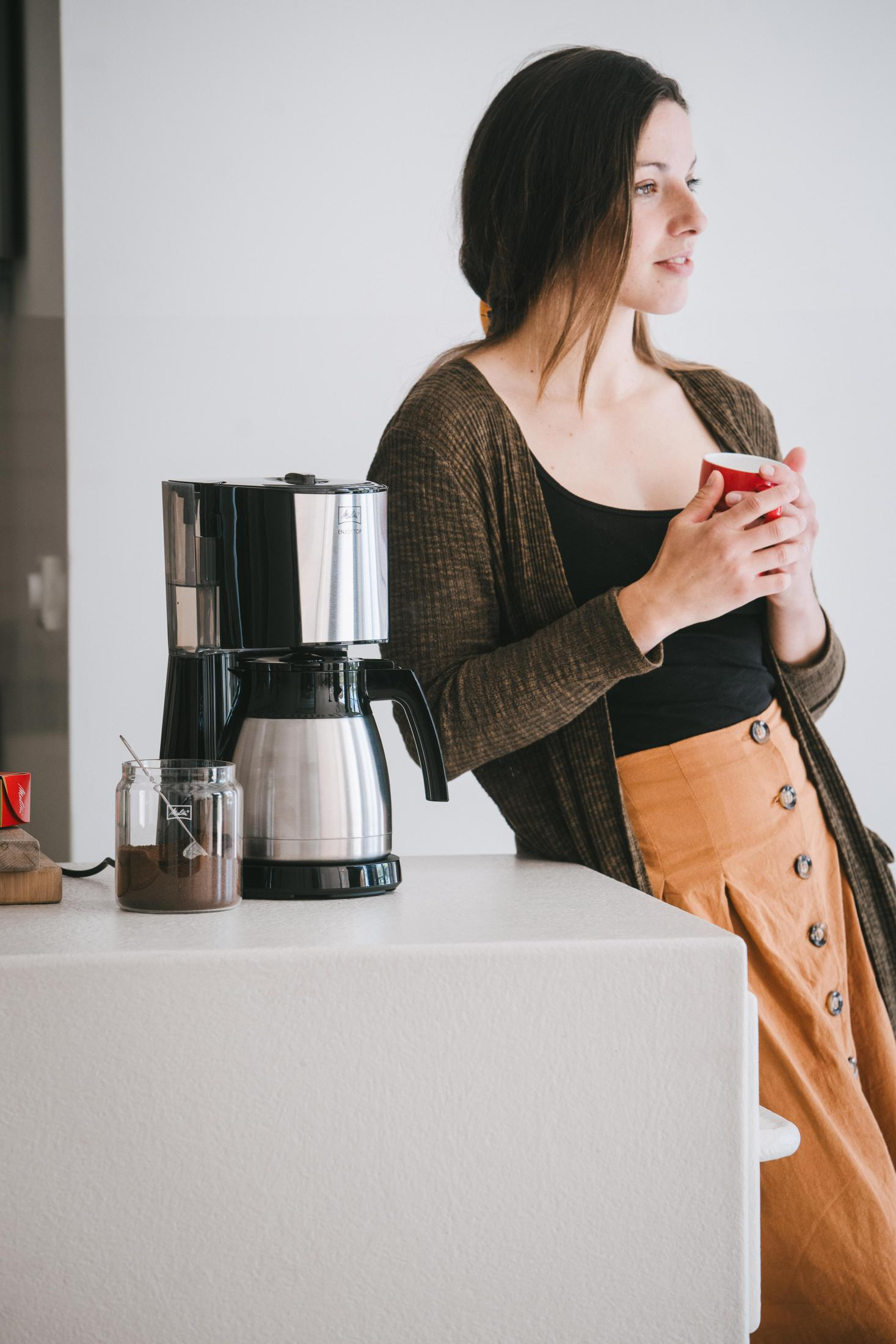 Kobieta trzymająca czerwony kubek obok ekspresu do kawy na białym blacie. Brązowy kardigan.