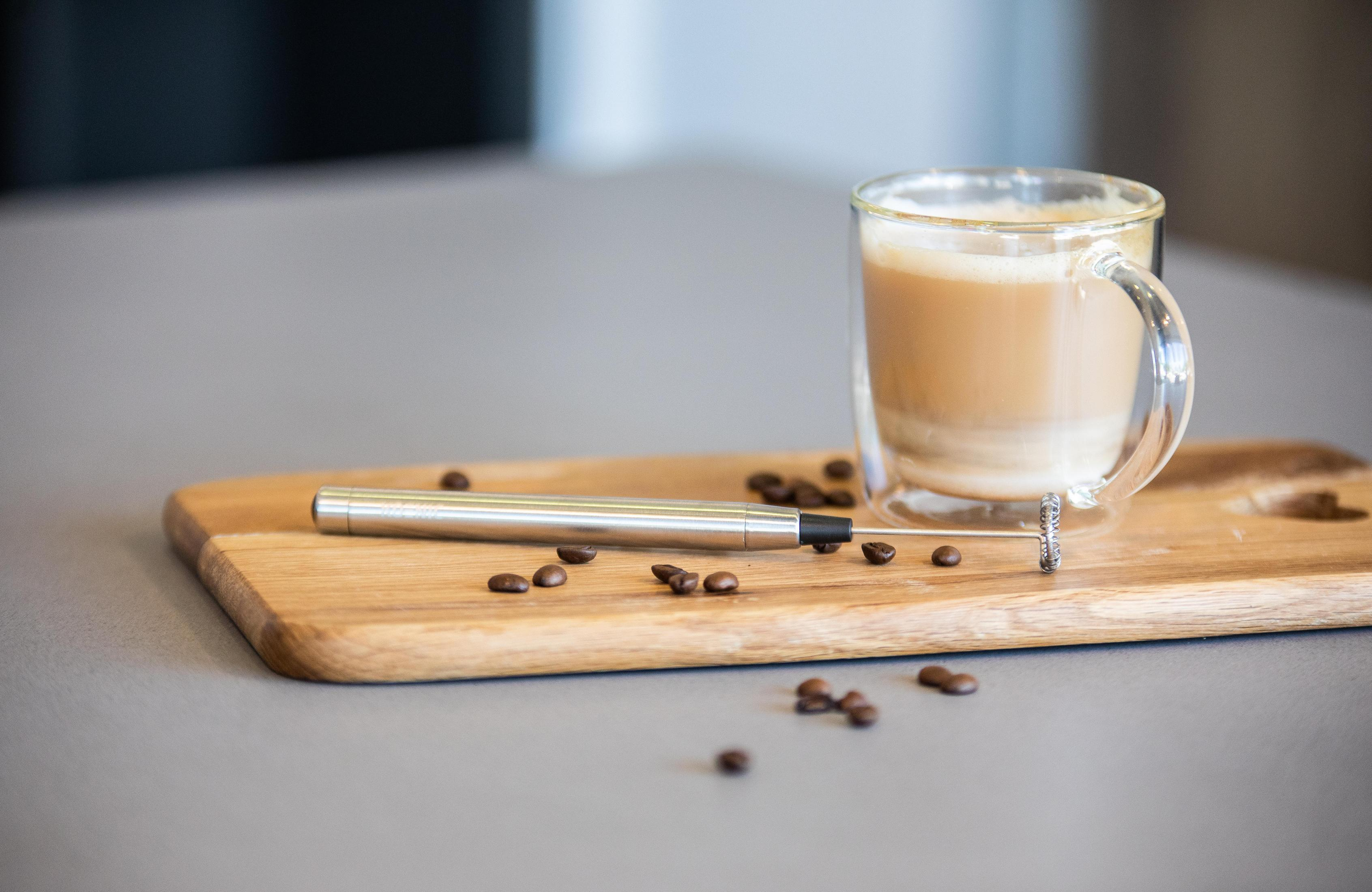 Ein Glasbecher mit Kaffee und Schaum auf einem Holzbrett mit einem Aufschäumwerkzeug und Kaffeebohnen.