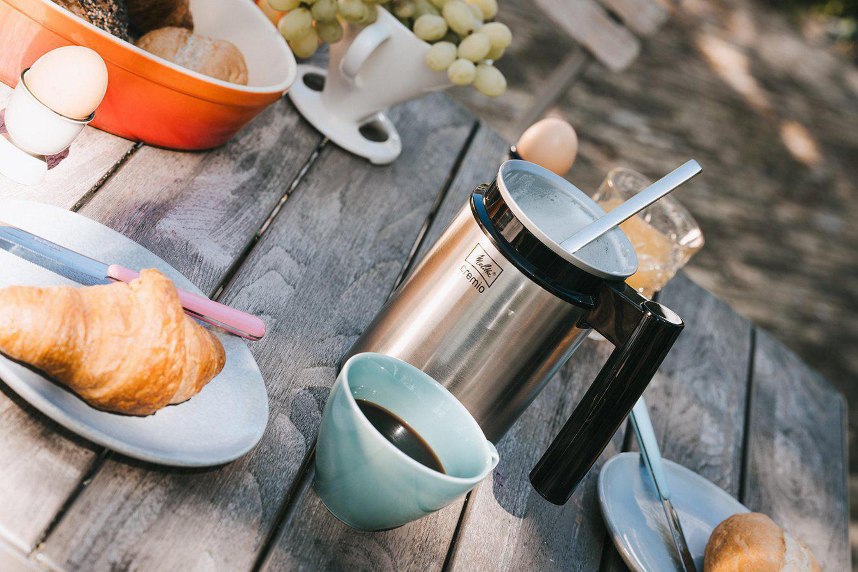 Śniadanie na drewnianym stole. Zawiera kawę, rogaliki, jajka i owoce.