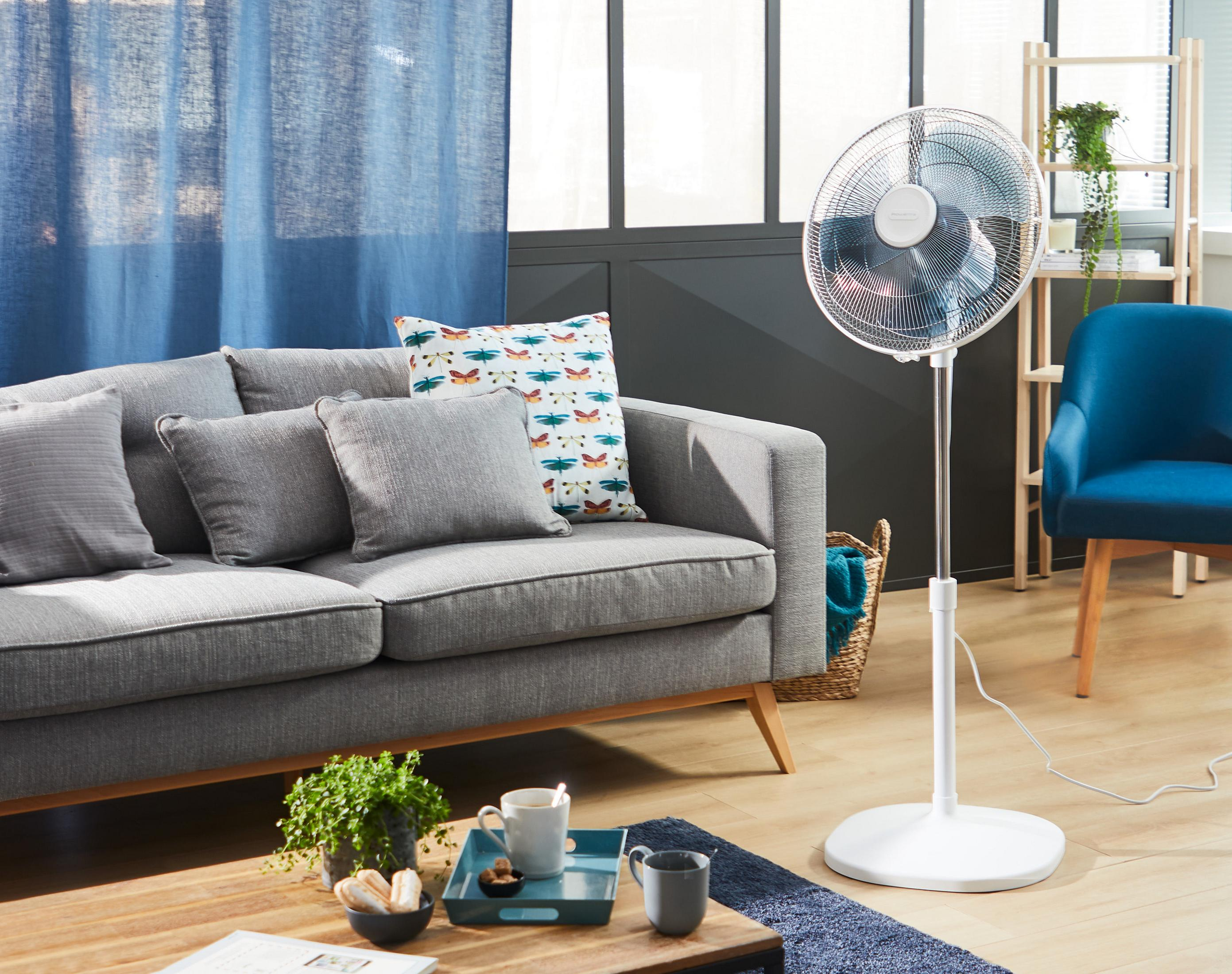 Salon z szarą sofą, poduszkami i wentylatorem. Widoczna niebieska zasłona i drewniany stół.