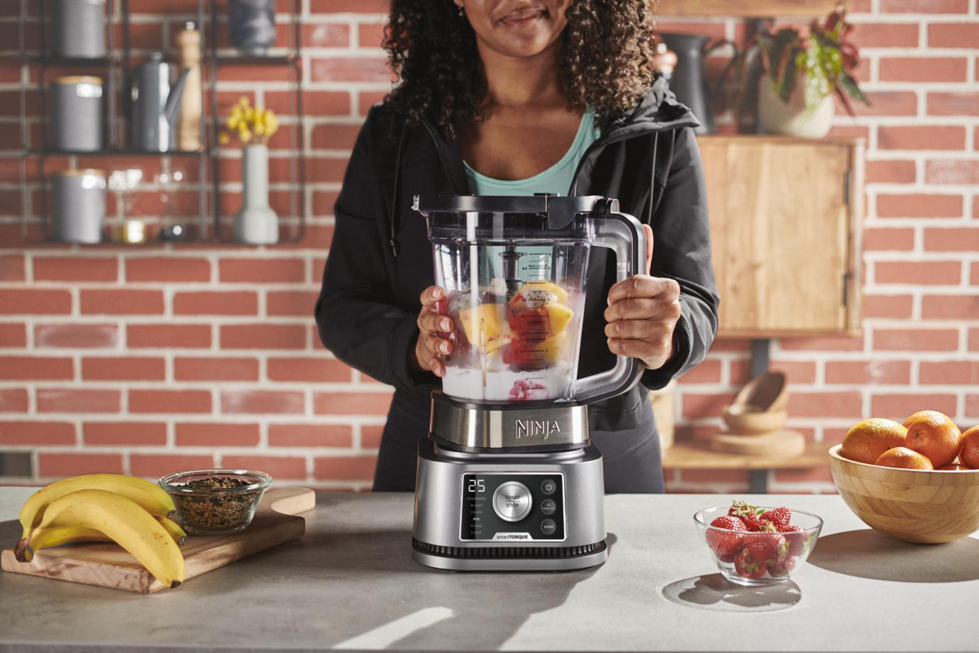 Woman using a Ninja blender, with various fruits and ingredients on a counter.