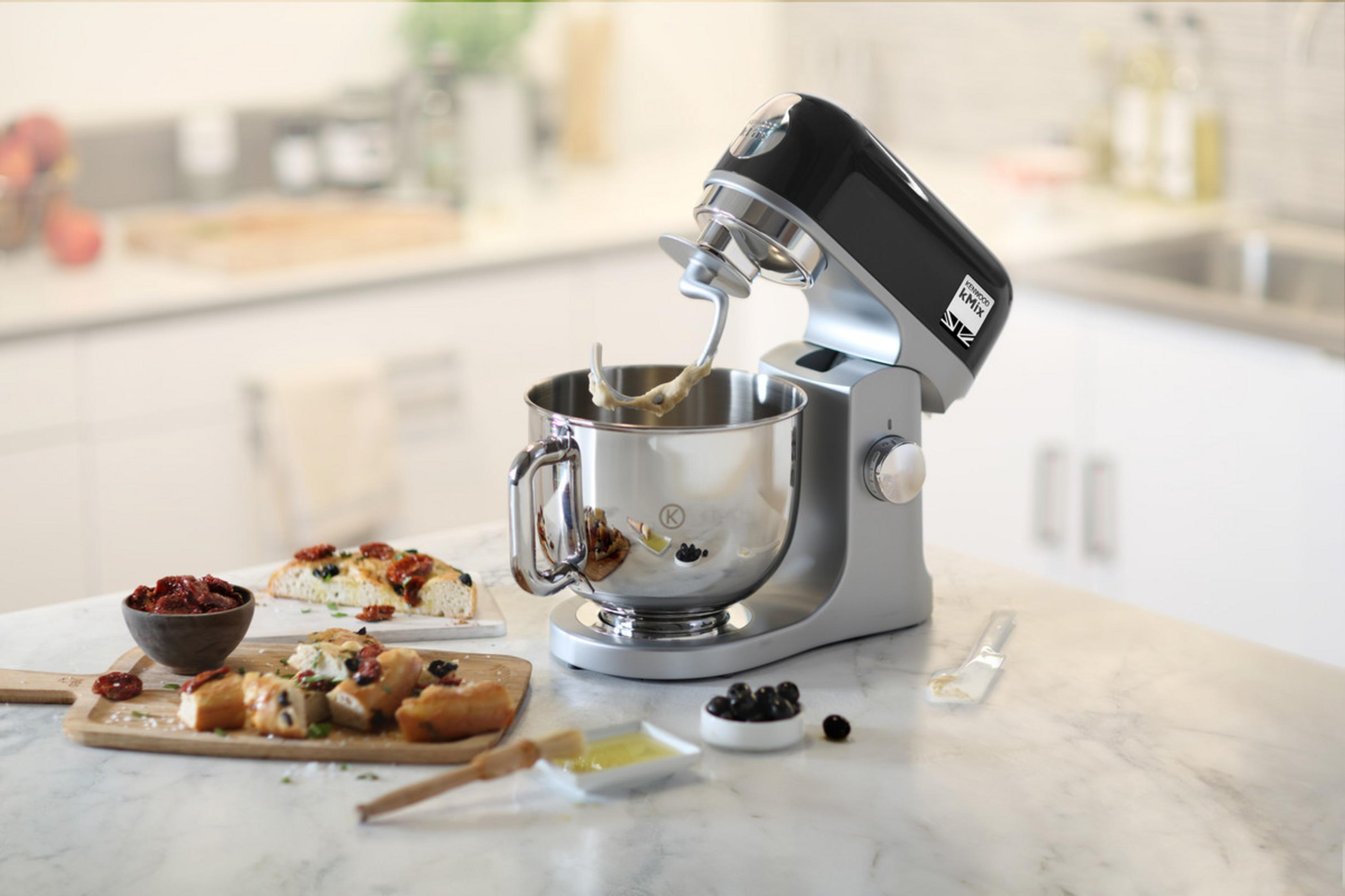 Ein silberner Küchenmixer mit einer Schüssel Teig und Focaccia, Oliven und Tomaten auf der Theke.