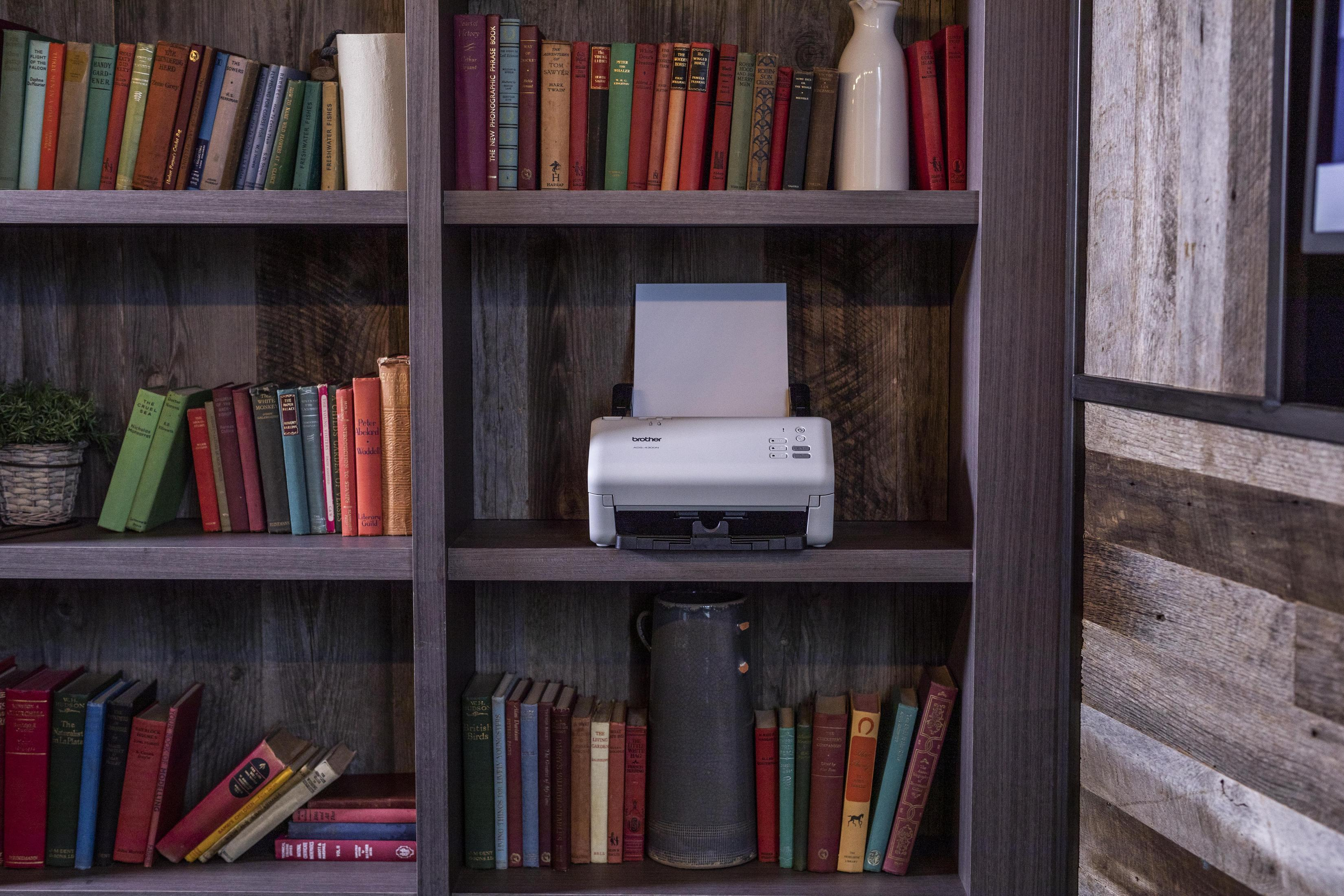 Drukarka na półce, otoczona książkami, w drewnianej biblioteczce. Drukarka jest biała z szarą górą.
