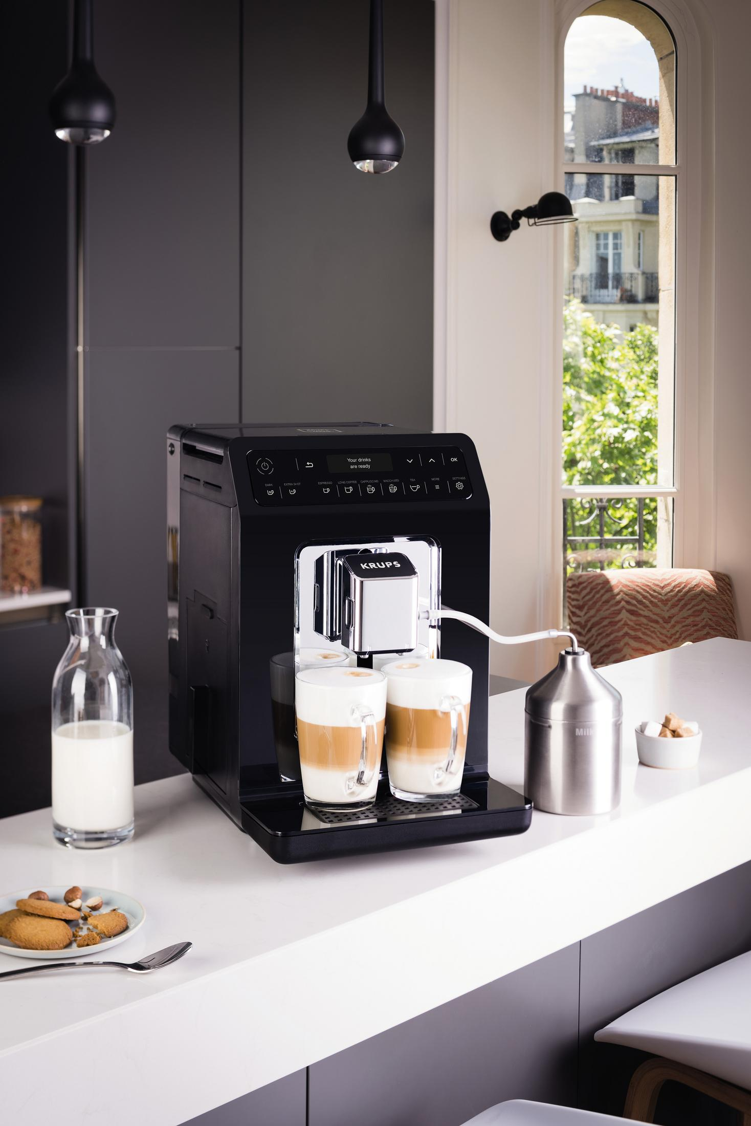 Ekspres do kawy z dwoma filiżankami latte. Na blacie butelka mleka, ciastka i cukier.