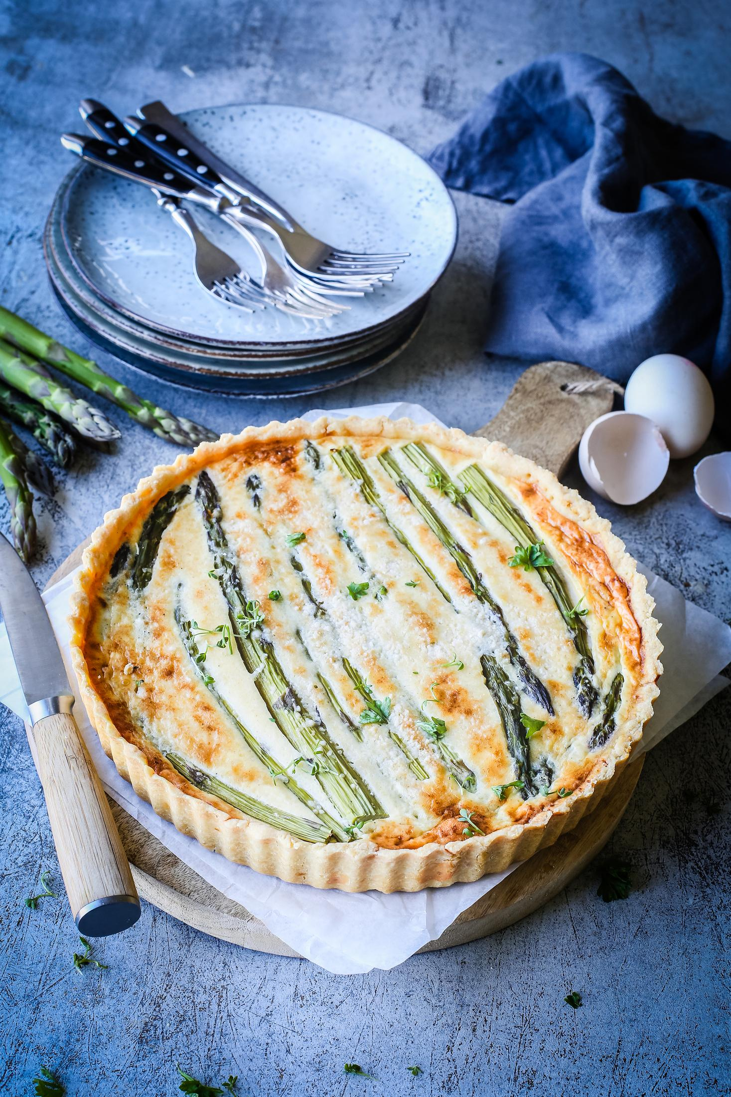 Quiche auf einem Holzbrett. Spargel ist die Hauptzutat, umgeben von Tellern und Besteck.