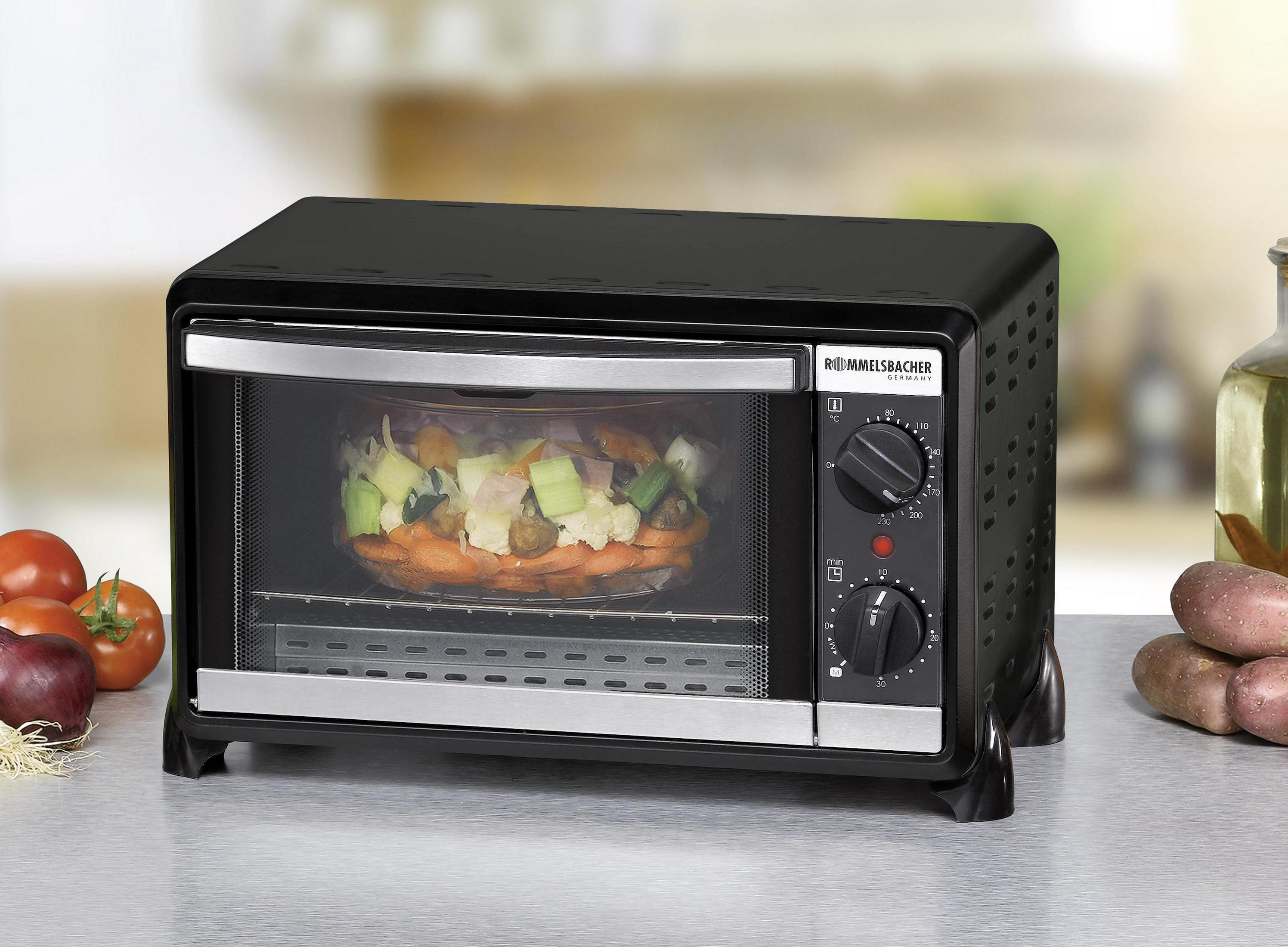 Czarny piekarnik na blacie z szklanymi drzwiami. Jedzenie widoczne w środku. Ziemniaki i pomidory w pobliżu.