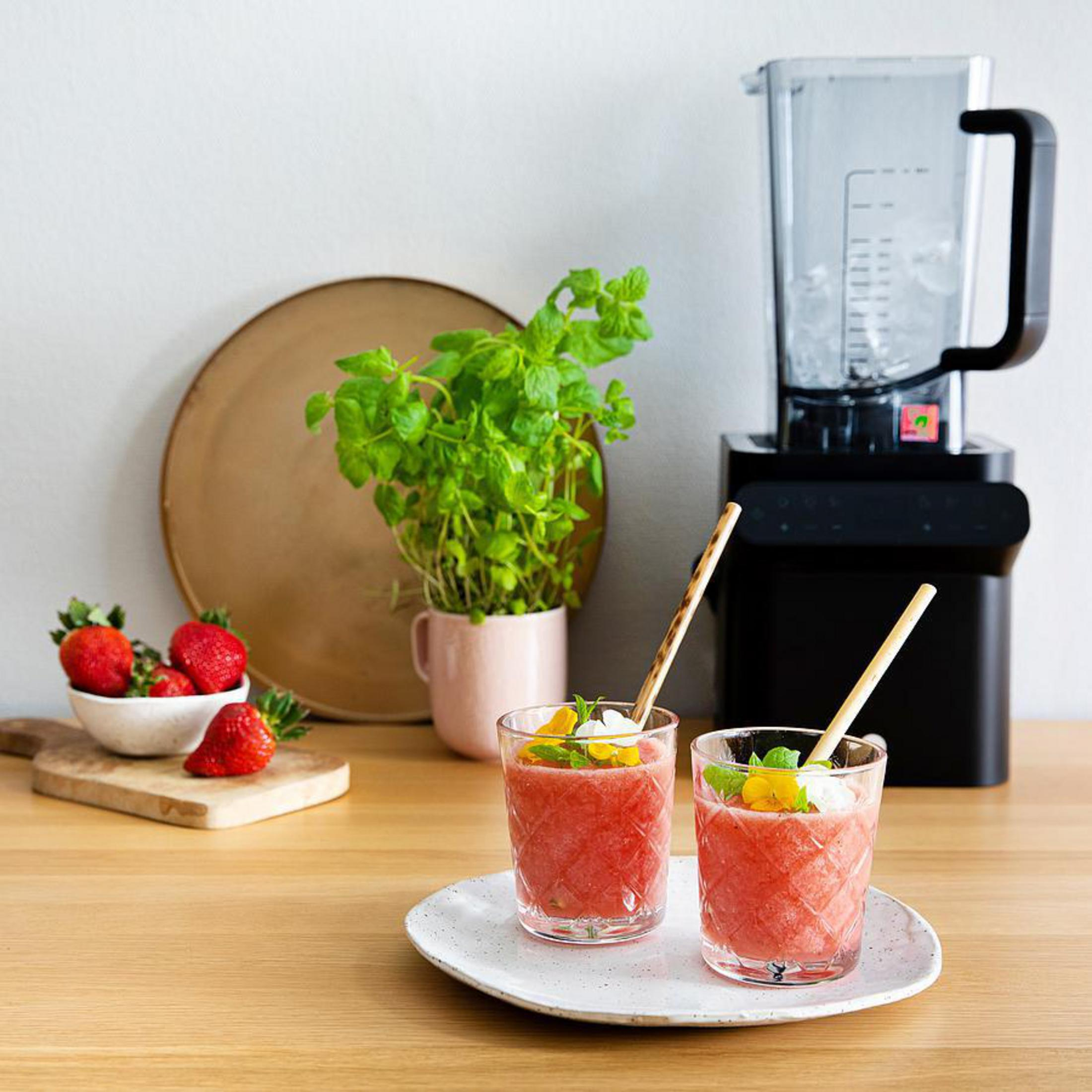 Zwei Gläser Smoothie mit Strohhalmen stehen auf einem Teller. Mixer und Zutaten daneben.