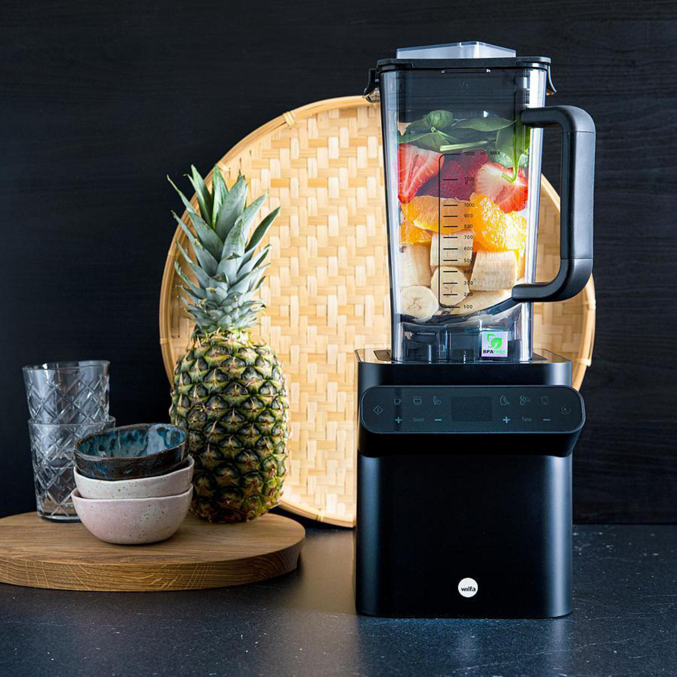 Ein Mixer mit Früchten, Ananas, Tablett und Schalen auf schwarzer Oberfläche.