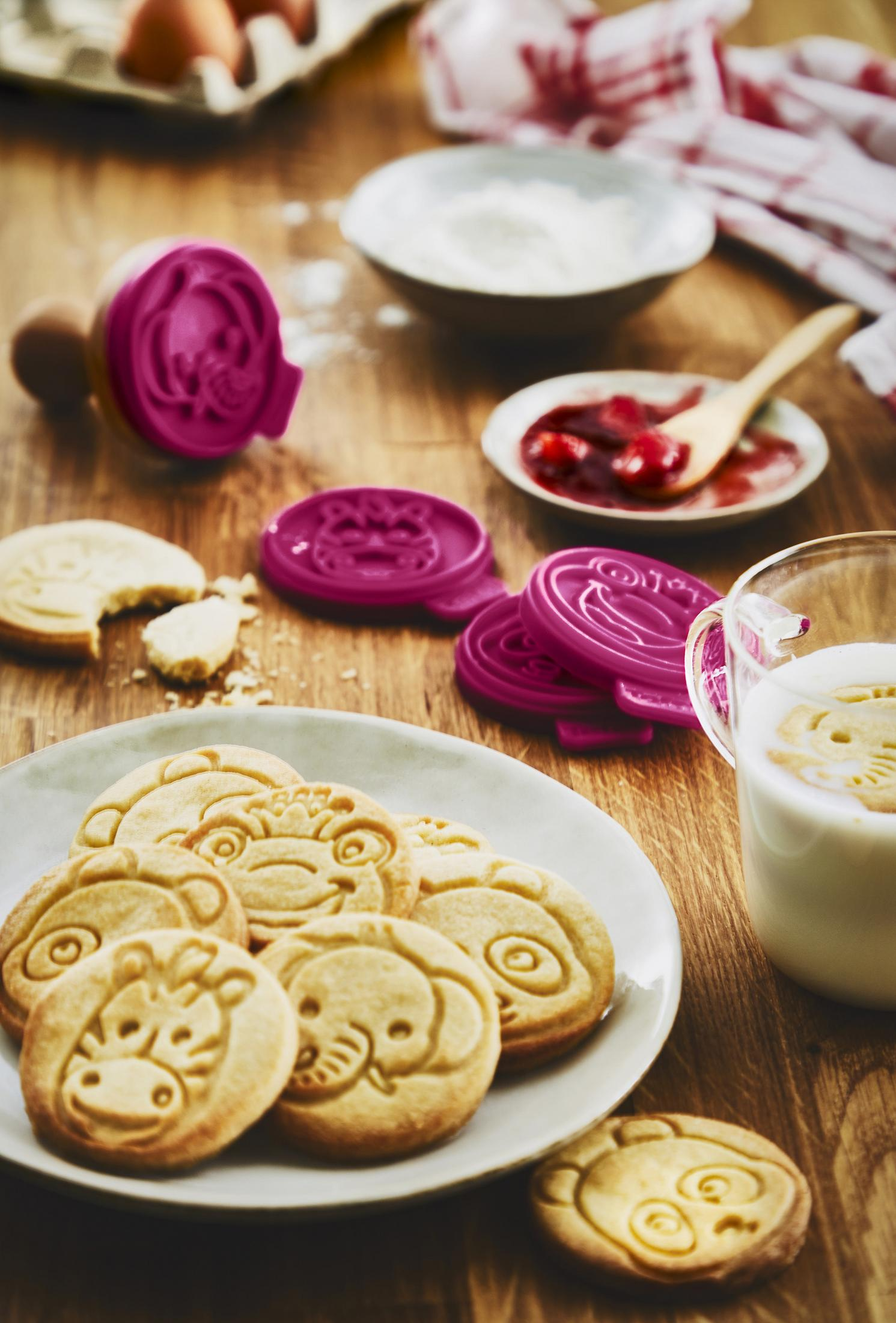 Biscotti a forma di animali su un piatto, formine, marmellata e latte su tavolo di legno.