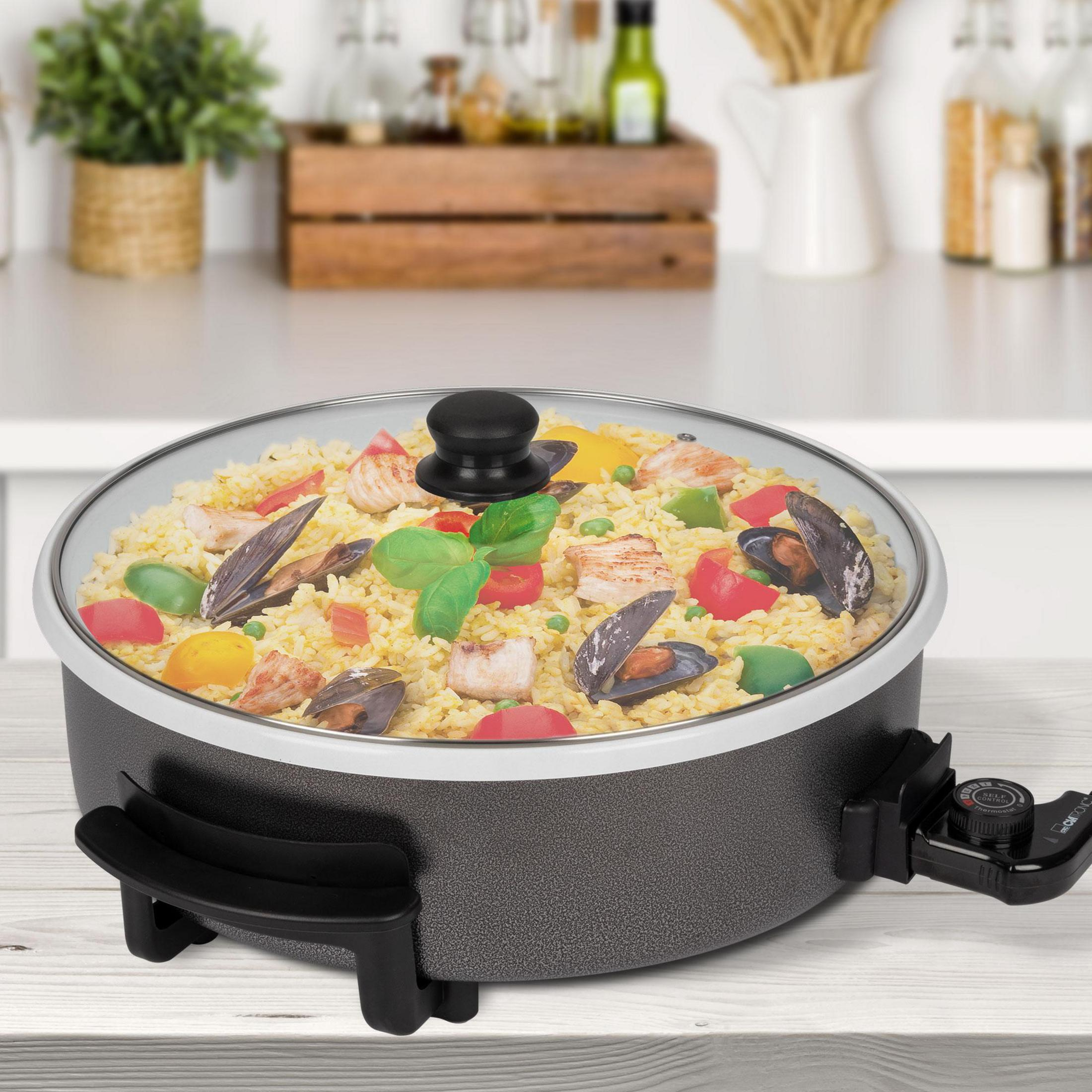 Elektryczna patelnia z ugotowanym jedzeniem pod szklaną pokrywką, na drewnianej powierzchni.