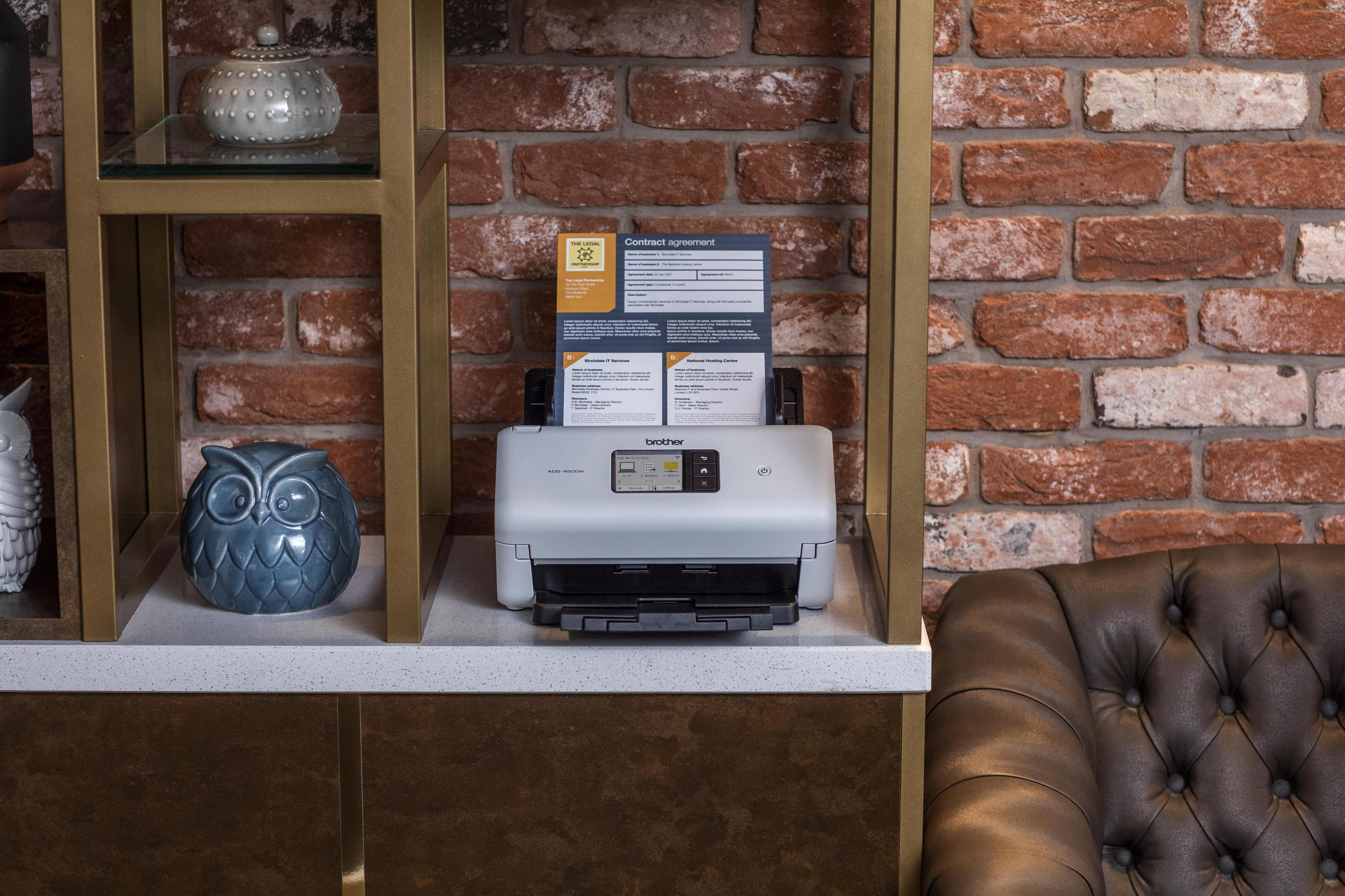 Drukarka stoi na półce. Obok niej niebieska sowa, a w tle ceglana ściana.
