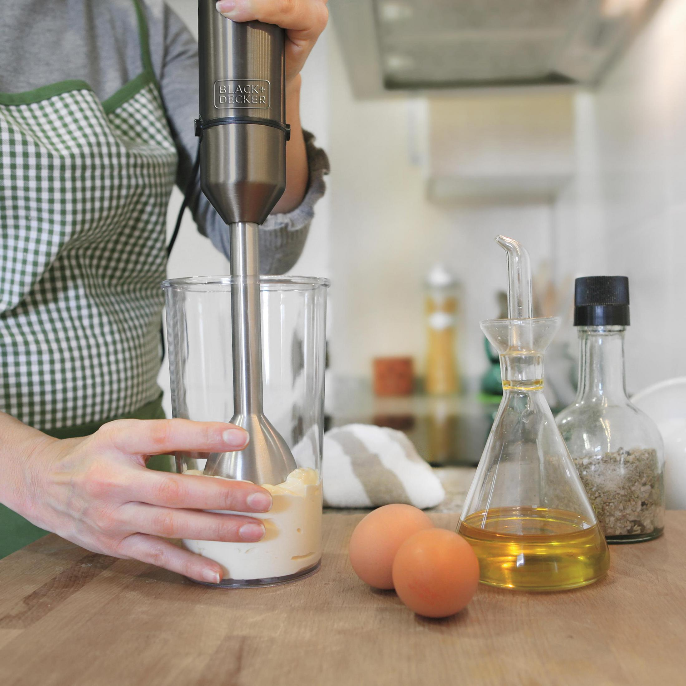 Persona che frulla ingredienti con un frullatore a immersione, con uova e olio nelle vicinanze.