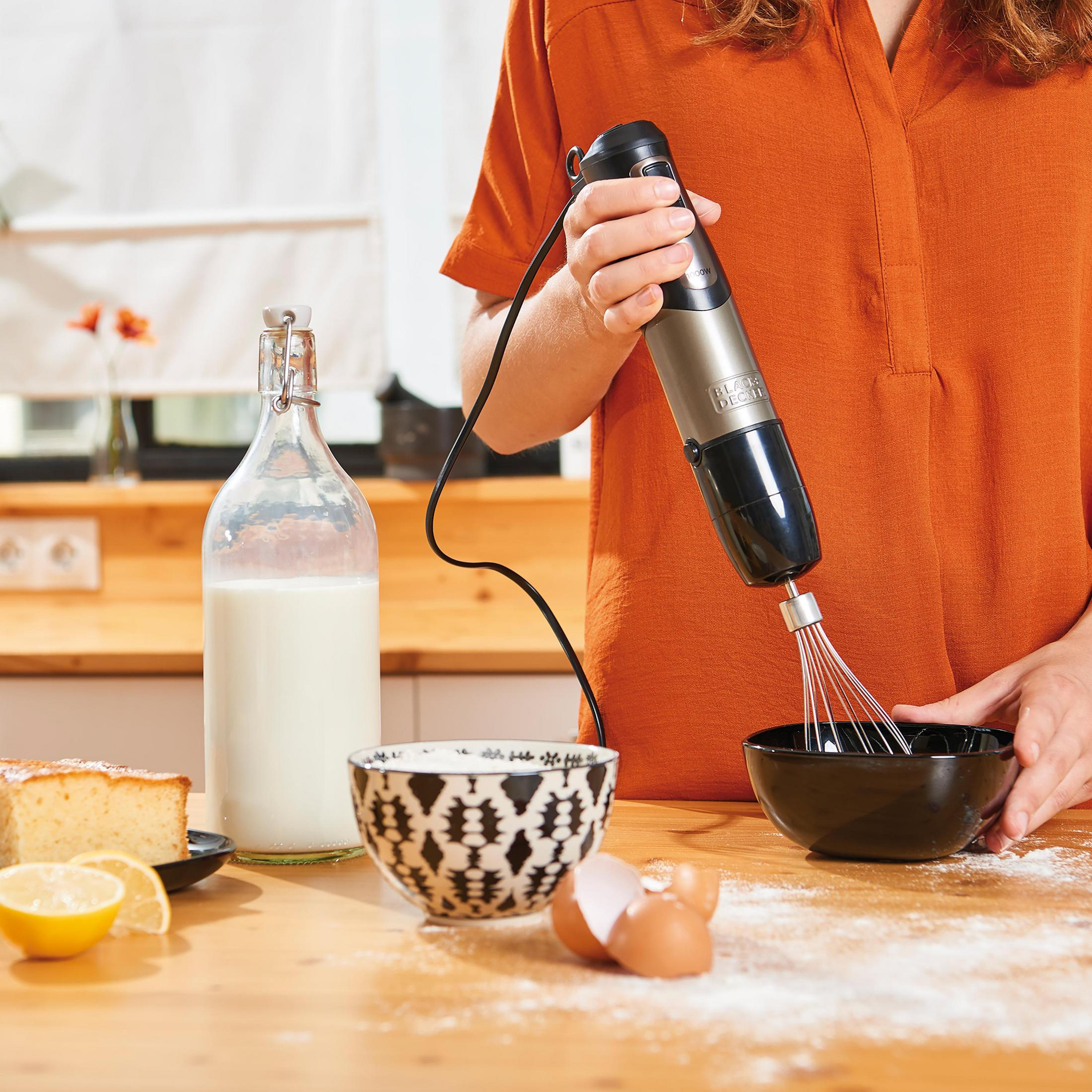 Una persona usa un frullatore a immersione per mescolare gli ingredienti in una ciotola nera. Sono visibili anche una bottiglia di latte e una torta.