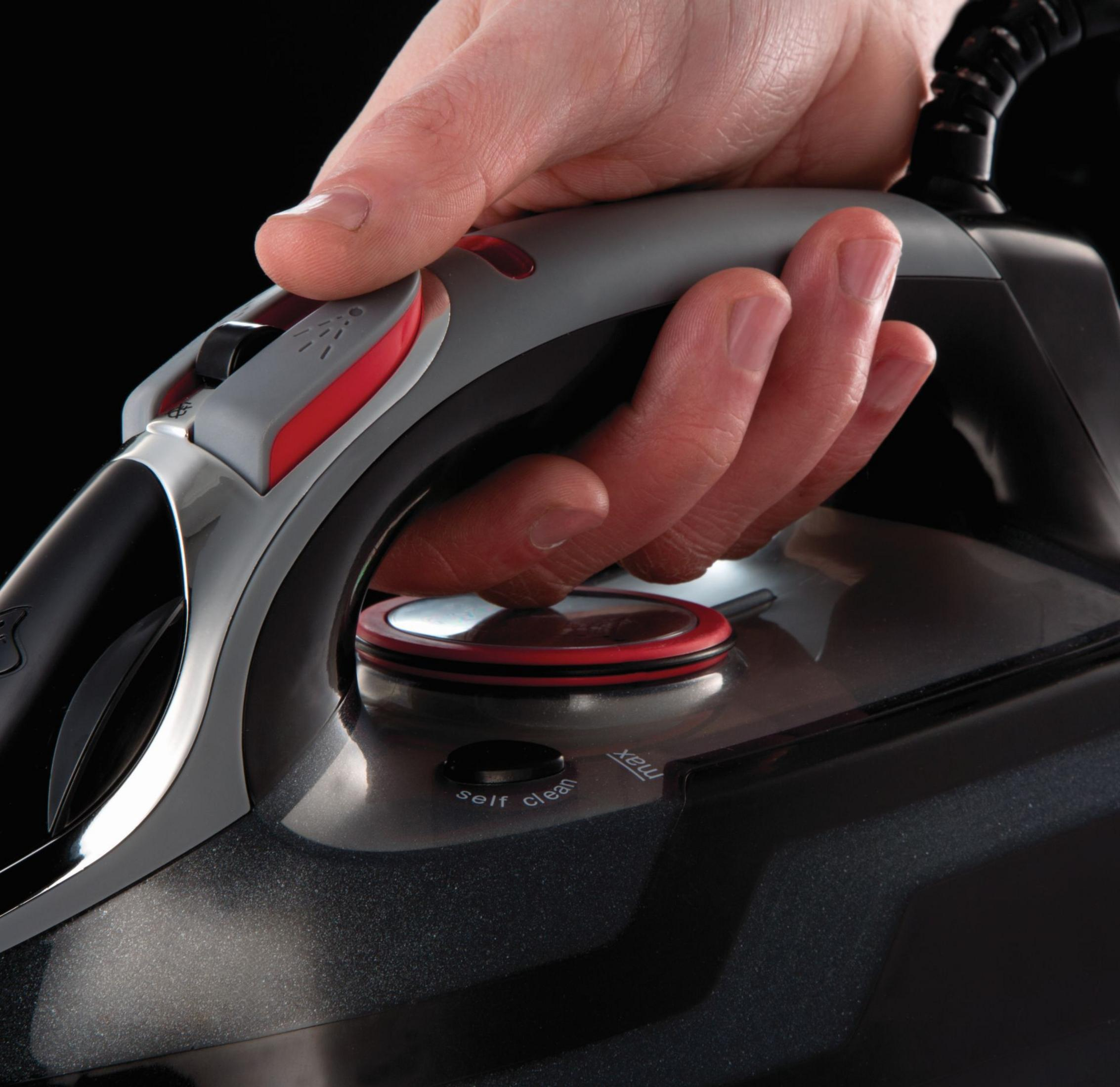 Ręka trzyma szare żelazko z czarnym tłem. Żelazko pokazuje funkcję samooczyszczania.