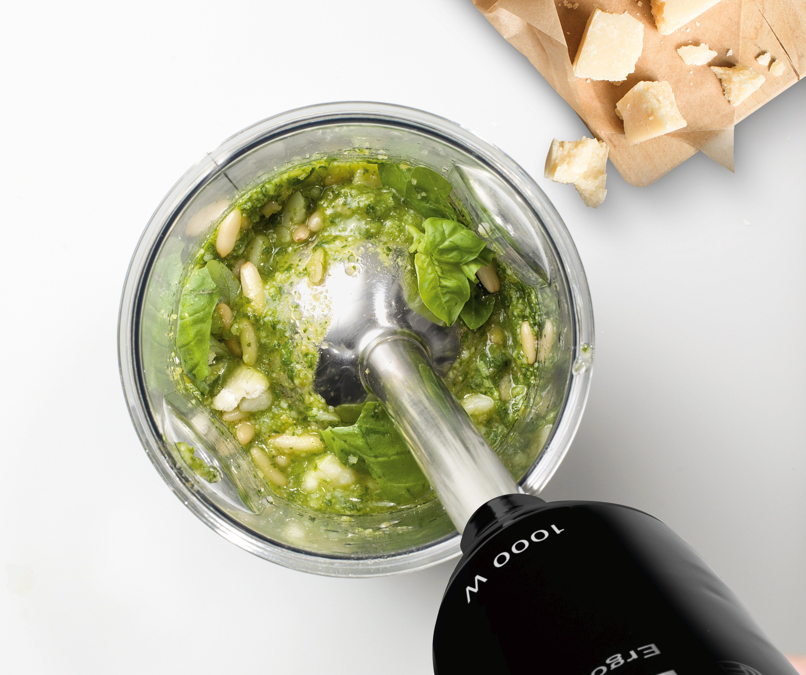 Een staafmixer in een glas met pesto-ingrediënten: basilicum, pijnboompitten en Parmezaanse kaas. Wit.