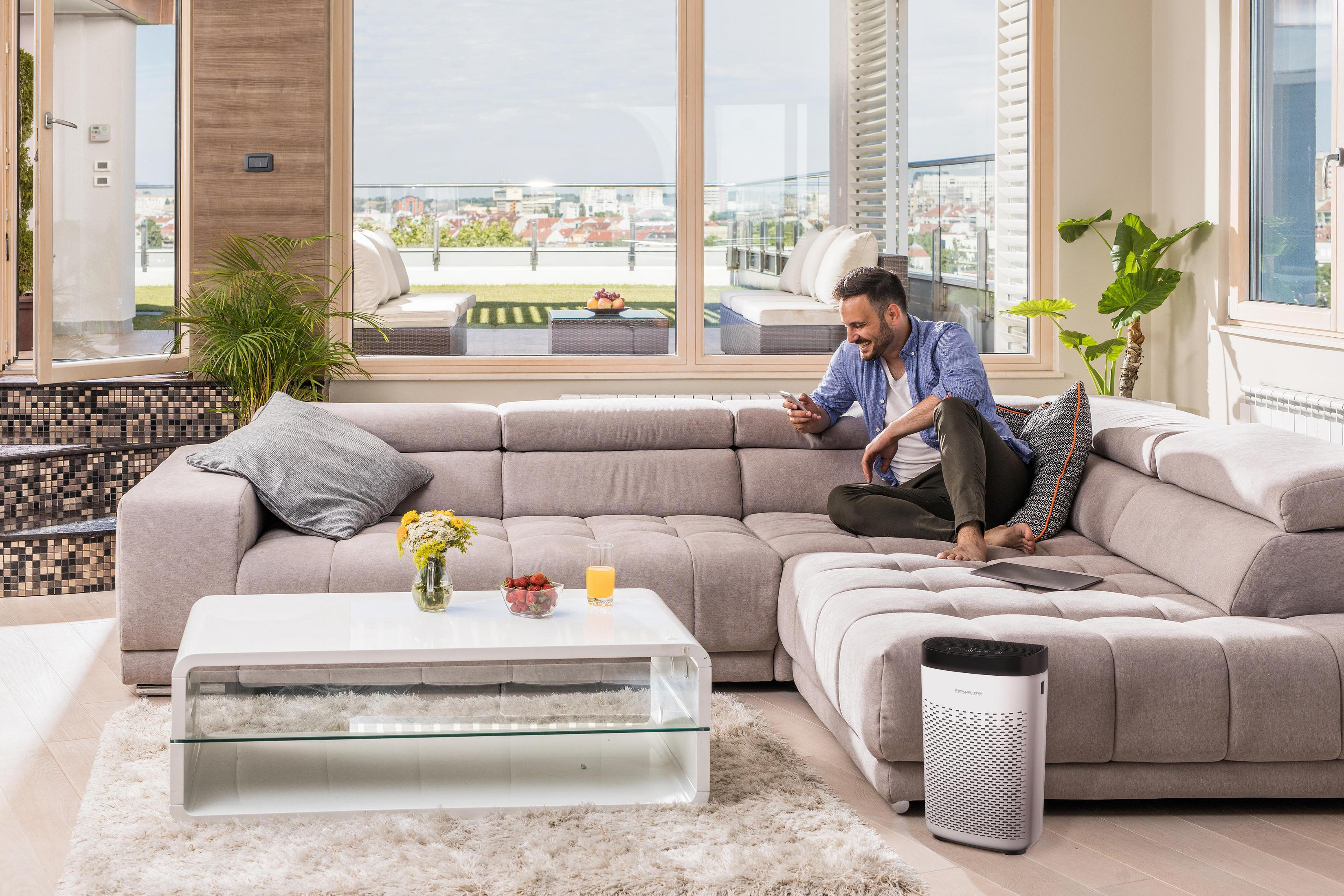Mann auf einem Sofa in einem modernen Wohnzimmer, der ein Telefon benutzt, mit einem Luftreiniger.