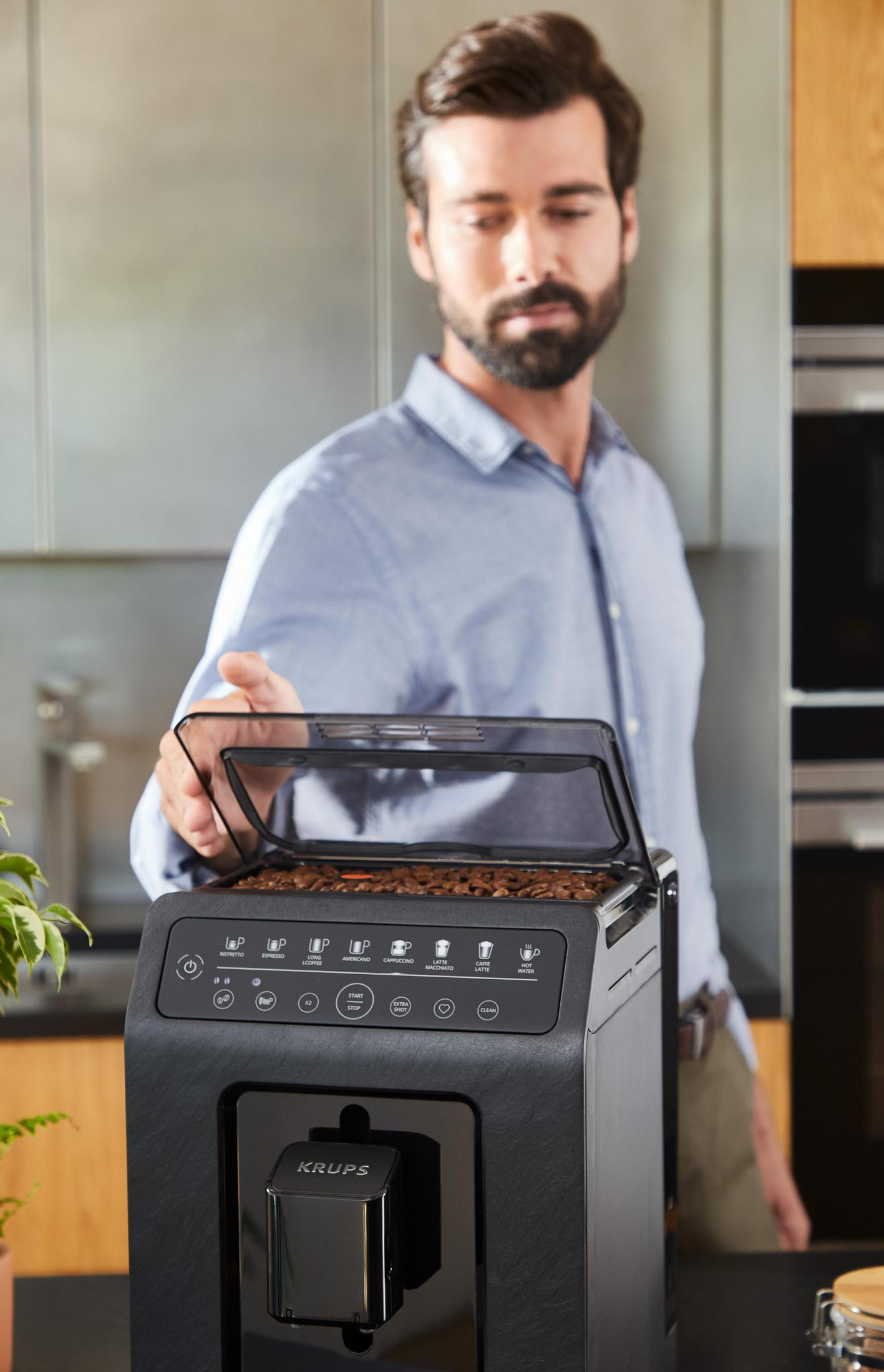 Mężczyzna otwiera pokrywę ekspresu do kawy, widoczne ziarna, tło kuchni.