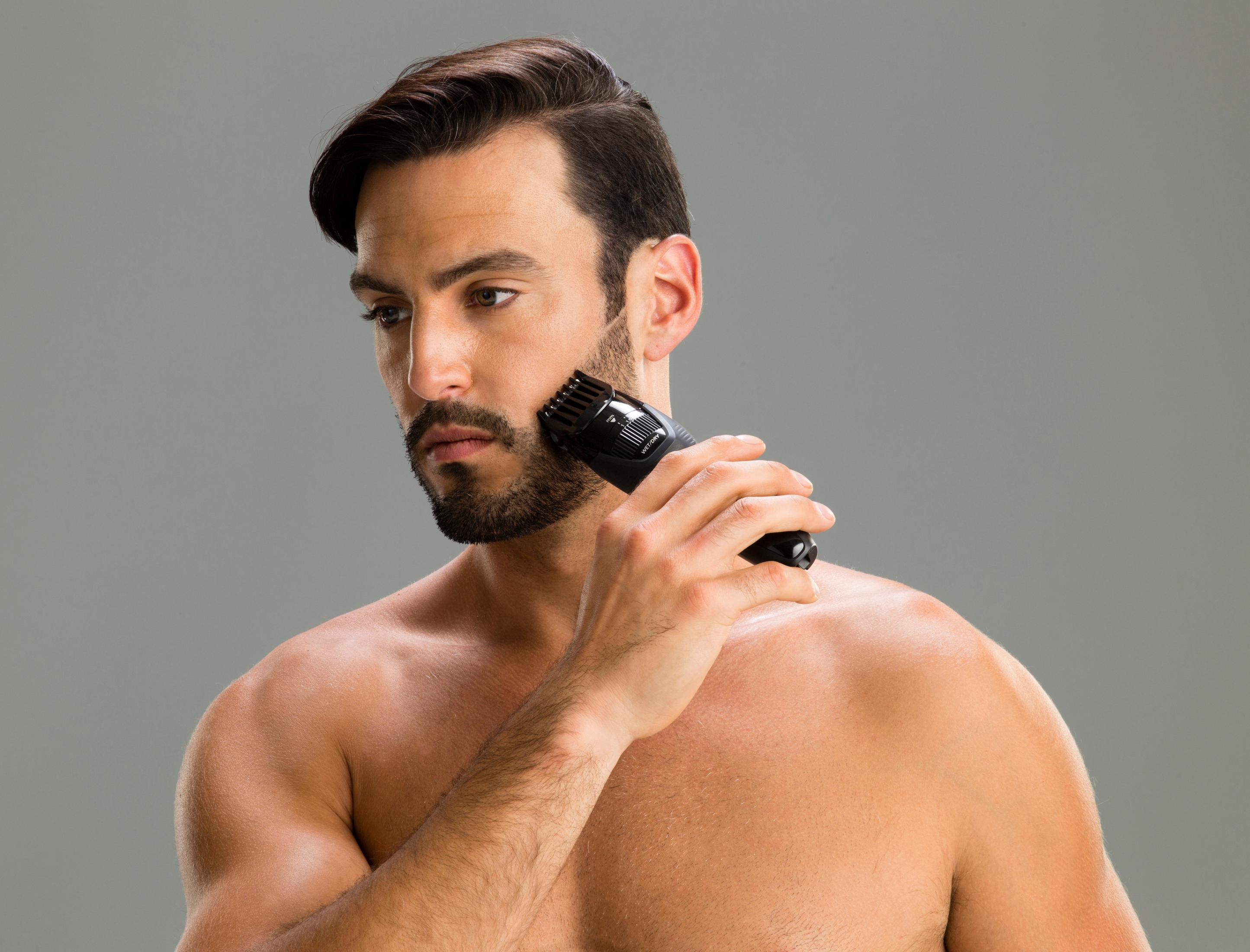 Un homme se rase la barbe avec une tondeuse noire. Il est torse nu.