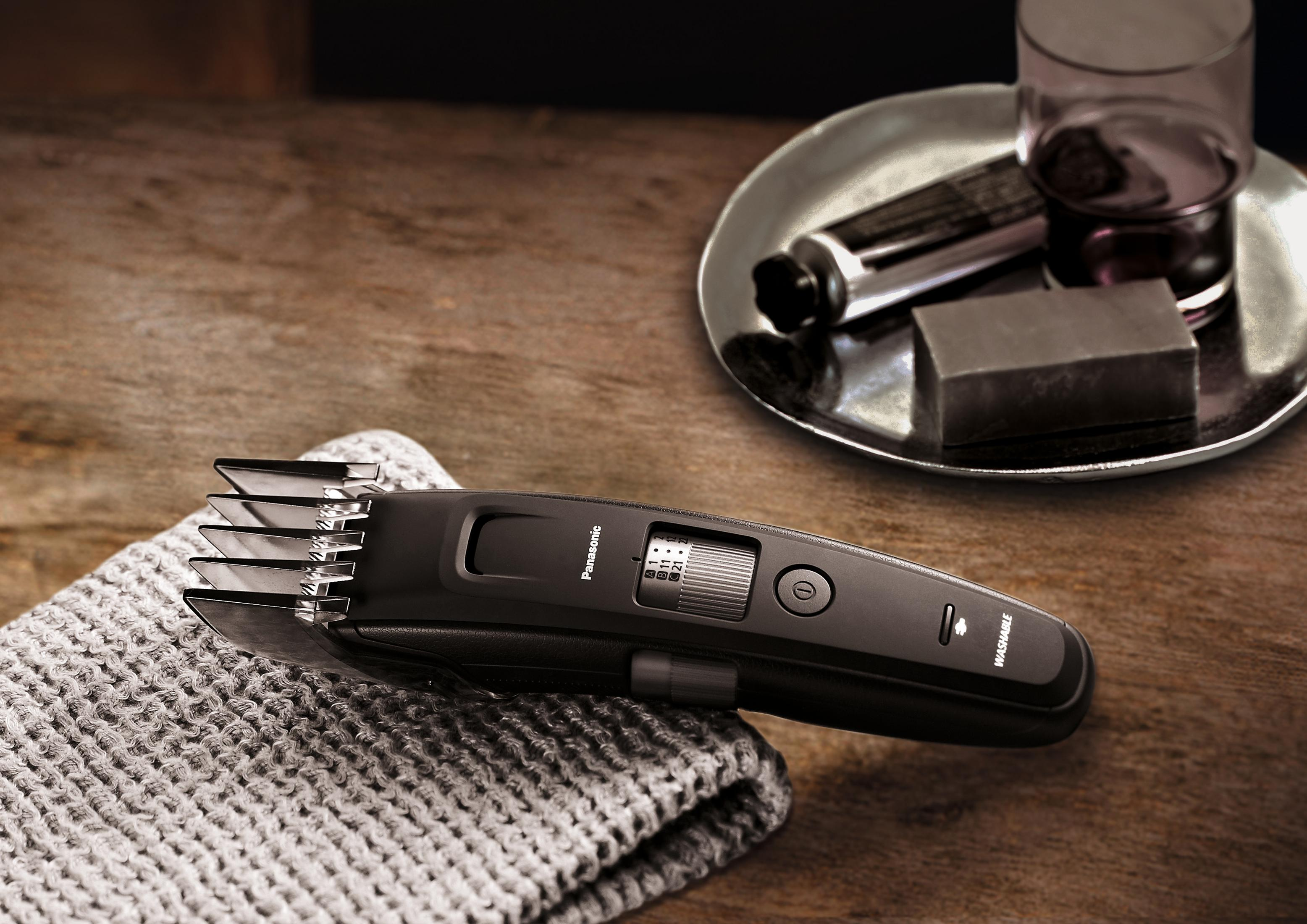 Schwarzer Haarschneider auf einem gefalteten grauen Handtuch, neben einem Glas und anderen Pflegeprodukten.