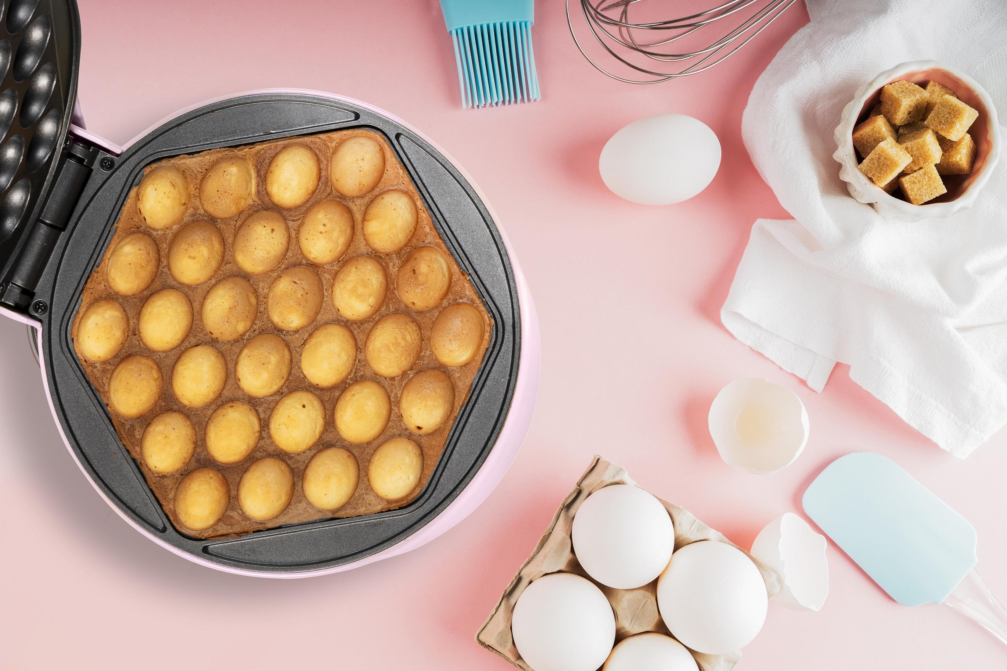 Macchina per waffle con waffle cotti, uova, zollette di zucchero e utensili da cucina su superficie rosa.