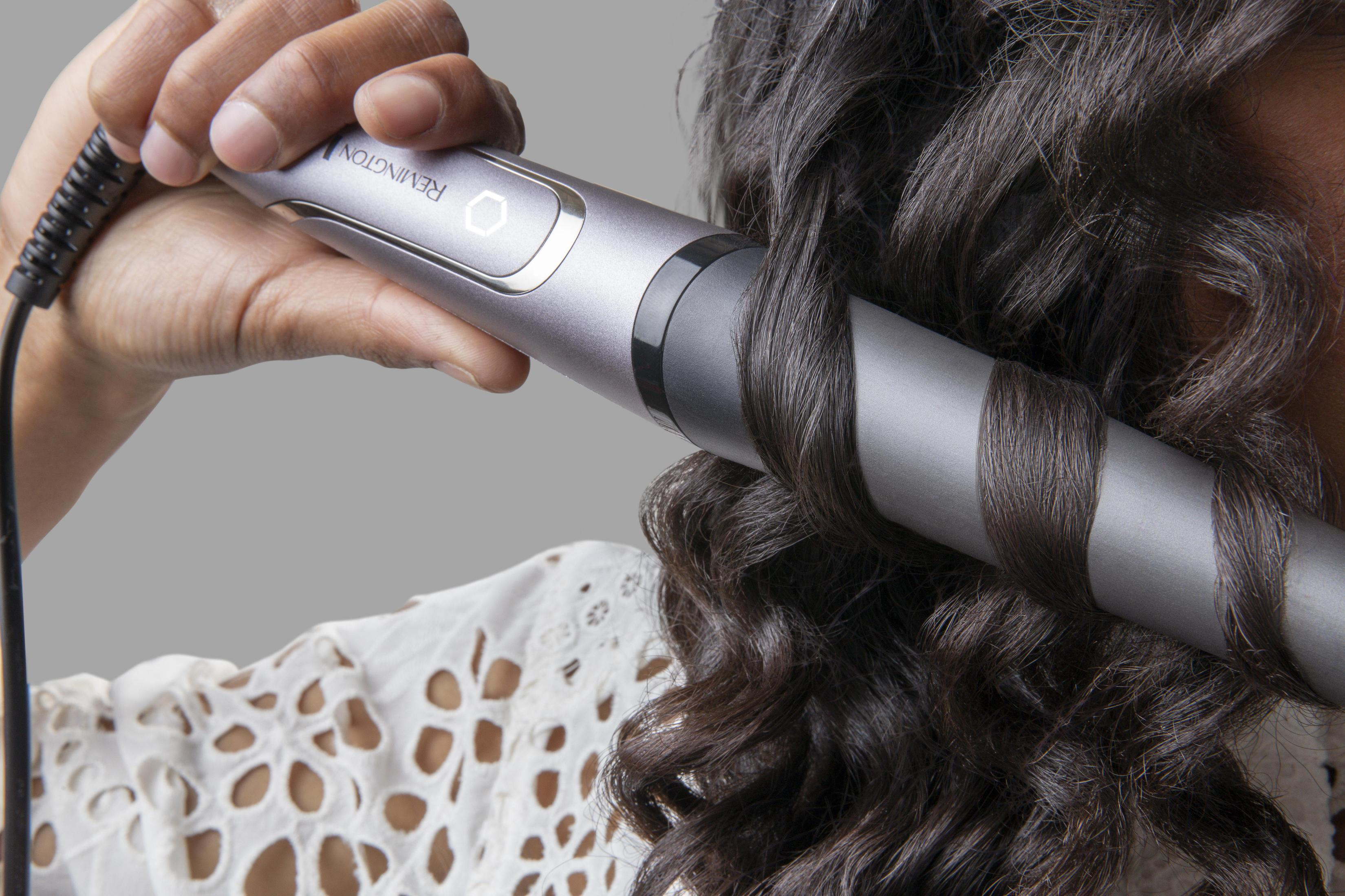 Osoba kręci włosy srebrną lokówką. Włosy są ciemne i falowane. Osoba ma na sobie biały top.