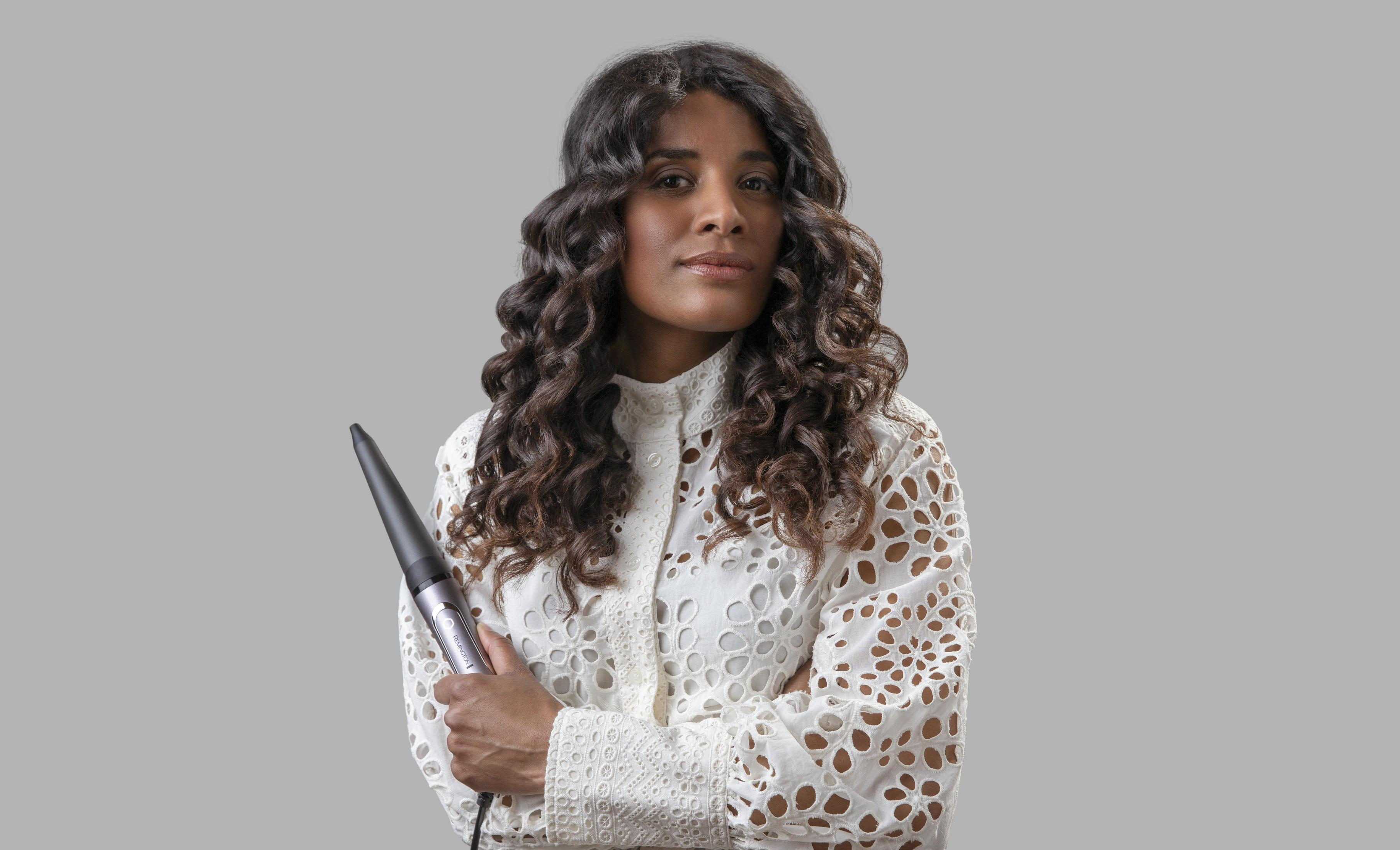 Kobieta z kręconymi włosami trzymająca lokówkę. Ma na sobie białą koronkową koszulę. Szare tło.