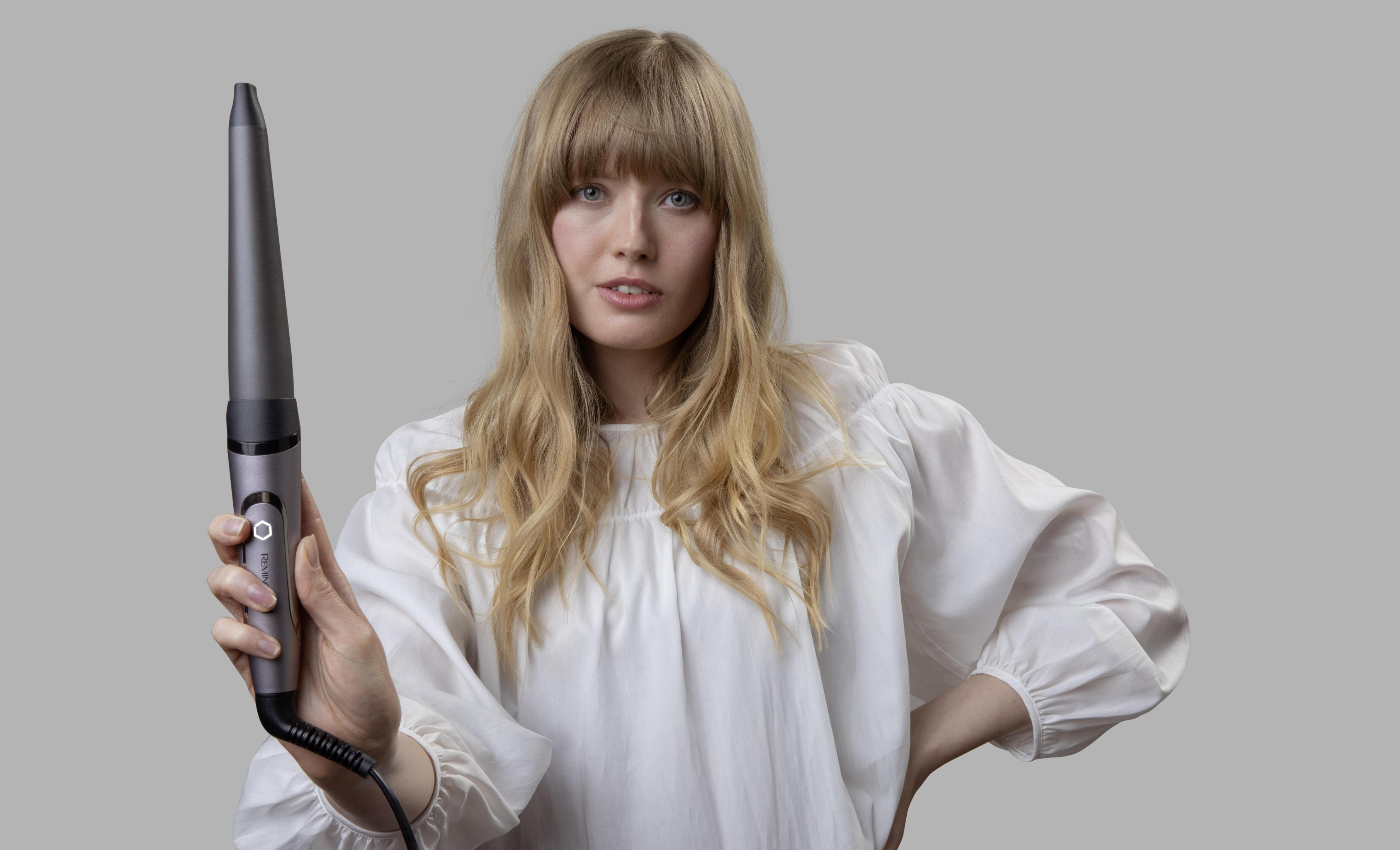 Blondynka trzyma szarą lokówkę, patrzy na widza. Ma na sobie białą koszulę.